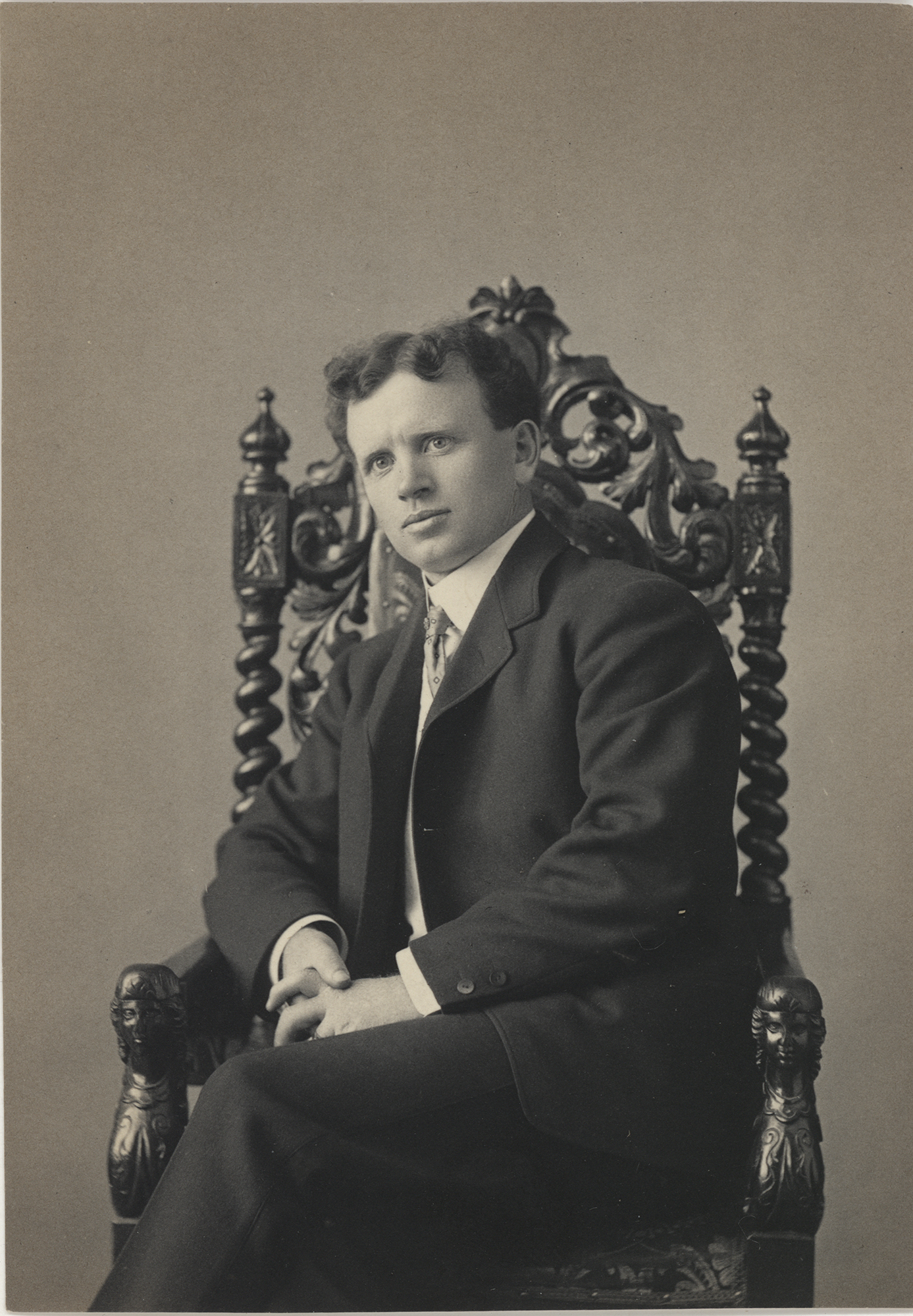 Photo of a man sitting in an ornately-carved wooden chair, posing for this portrait taken around 1900. The young man is dressed in a dark colored suit, with a white dress shirt and a necktie. He has short dark hair that has tamed curls on the top of his head. He is sitting in the chair with his left leg crossed over the right, and this hands clasped in a relaxed manner, resting his right elbow on the arm of the chair. His suit jacket is unbuttoned. The background is plain behind him.