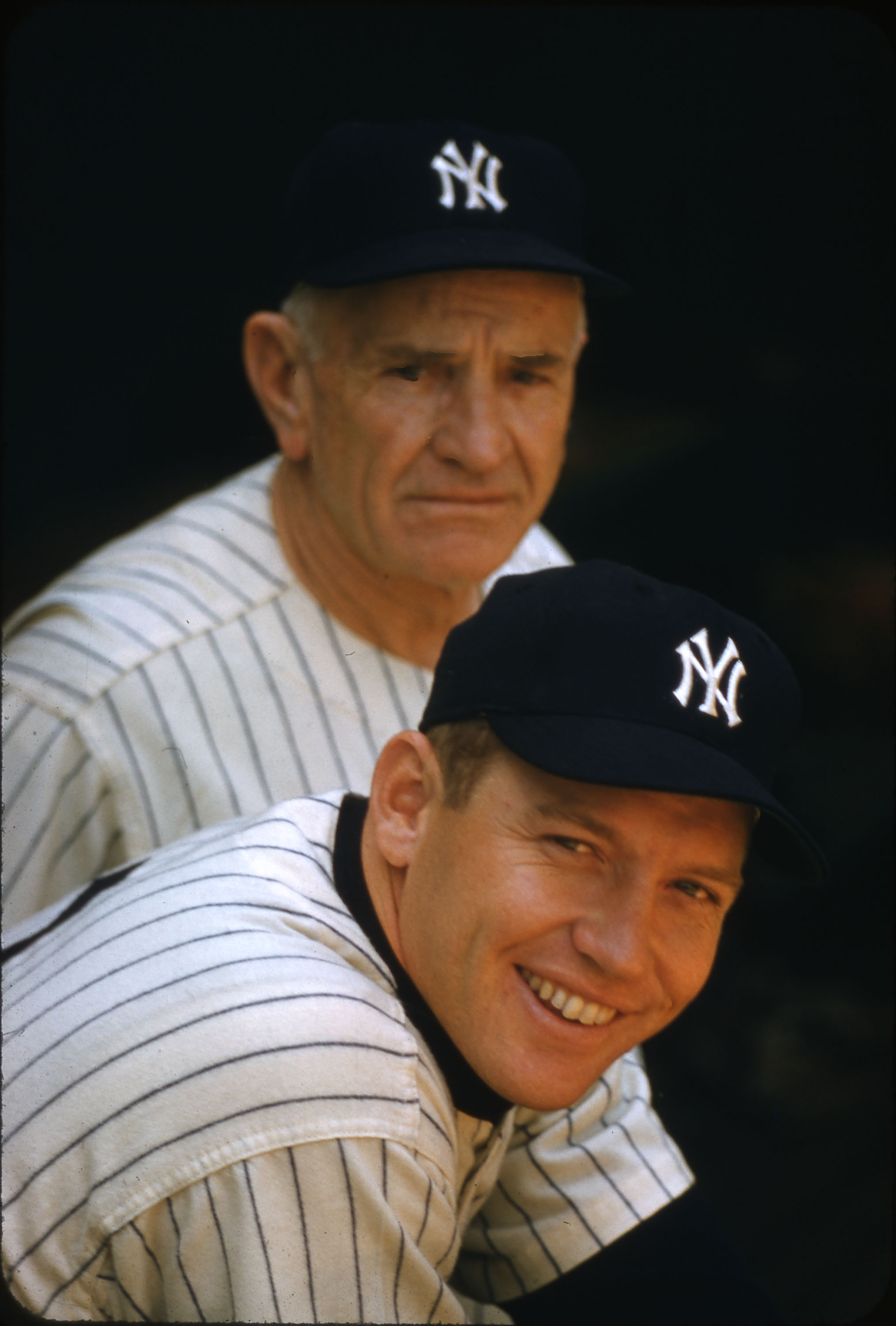 Photo of baseball player Mickey Mantle, looking at the camera and smiling with a wide, bright smile. Another man stands behind him, looking at him smiling to the camera. They both wear Yankees uniforms with dark colored caps with white Yankees logos, and white tops with dark, thin vertical pinstripes. 