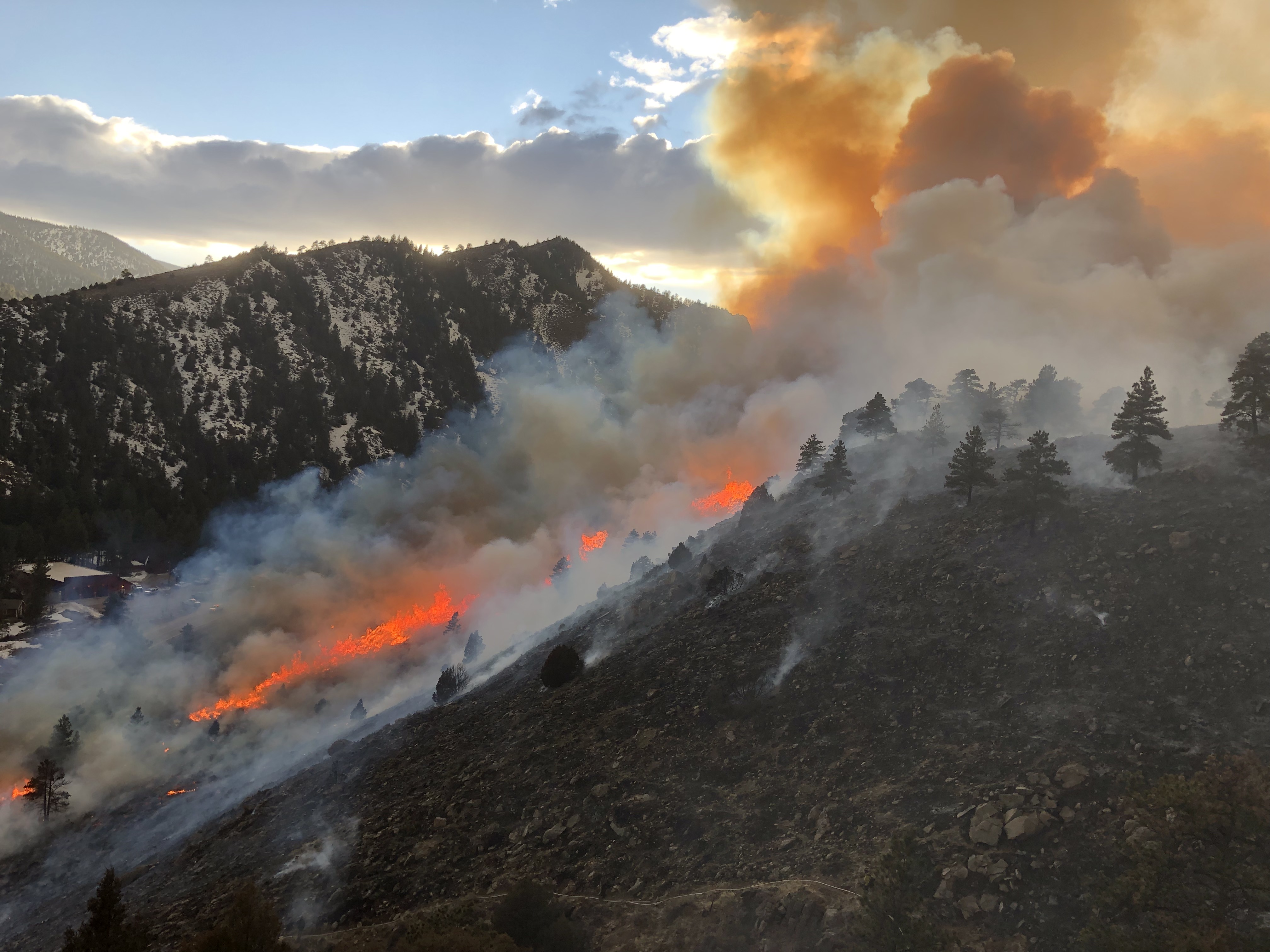 Photo of a wildfire burning vegetation up the side of a hill. The vibrant red and orange flames are consuming trees and releasing clouds of smoke into the sky, which are colored bright orange as they are backlit by the sun. The sky above the mountains is a bright blue, with a few atmospheric clouds present.