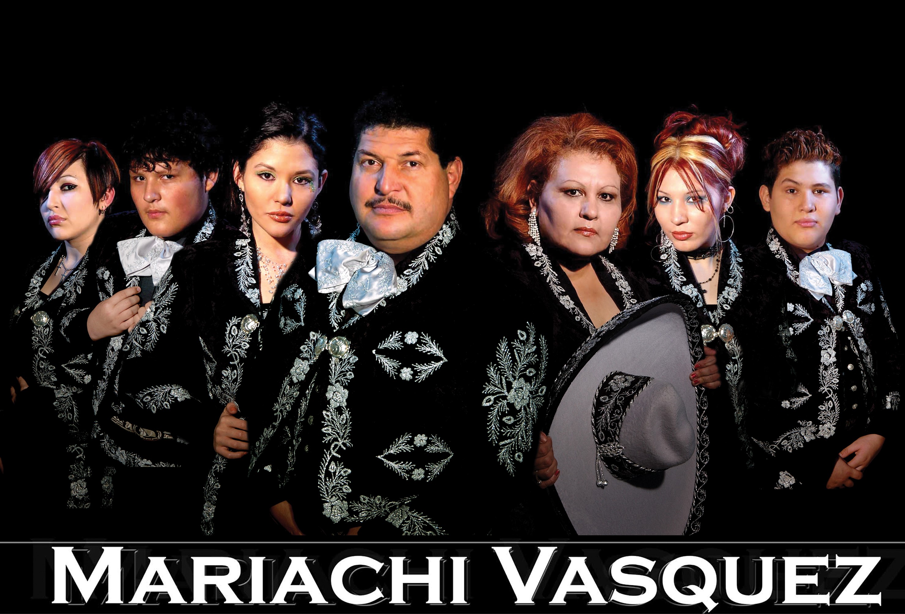 Photo of a group of 7 members of Mariachi Vasquez, as noted at the bottom of the image. The members of the group are wearing ornate black Mariachi outfits, embellished with embroidered white flowers on the collars, sleeves, on the jacket fronts and along the plackets. The males also wear a large white bow tie, one of the women holds a white sombrero trimmed in black. The background of the photo is all black.