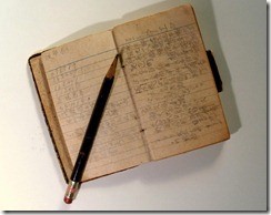 Photo of a small pocket-sized journal, about as tall as a pencil. The pages are filled with handwriting, scribed in Japanese characters. A pencil lays across the pages of the book.