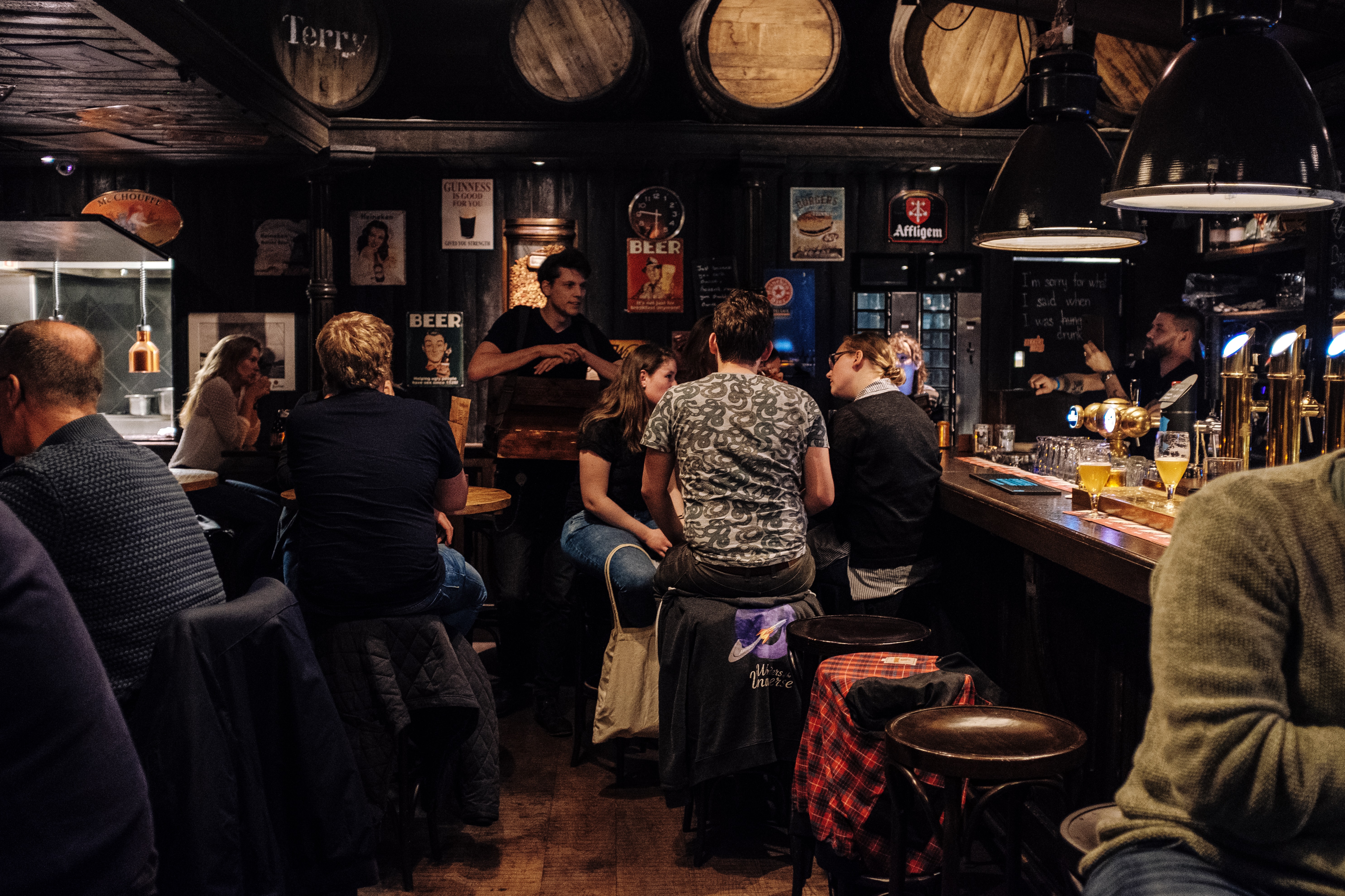 Photo of people sitting in a bar, chatting with one another. Their coats are draped over the barstools upon which patrons are sitting. The bar itself is on the right, with the bartender using a tap to pour a beer. The walls are covered in dark wood paneling with signs advertising different beers like Guinness. Above the seating area, wooden barrels are used as decoration.