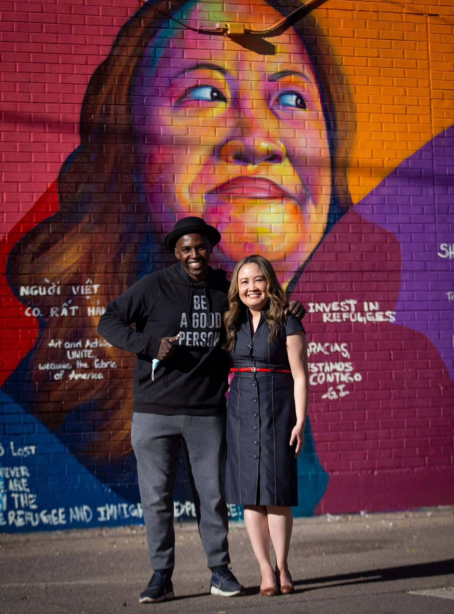 Photo of Nga Vương-Sandoval with Artist/Muralist Thomas Evans (I Am Detour). They are standing with arms around each other in front of the mural Evans created to recognize Vuong-Sandoval's advocacy work. The mural is an exterior brick wall, and the close up image of her face has been created using bright colors of red, orange, and pink.