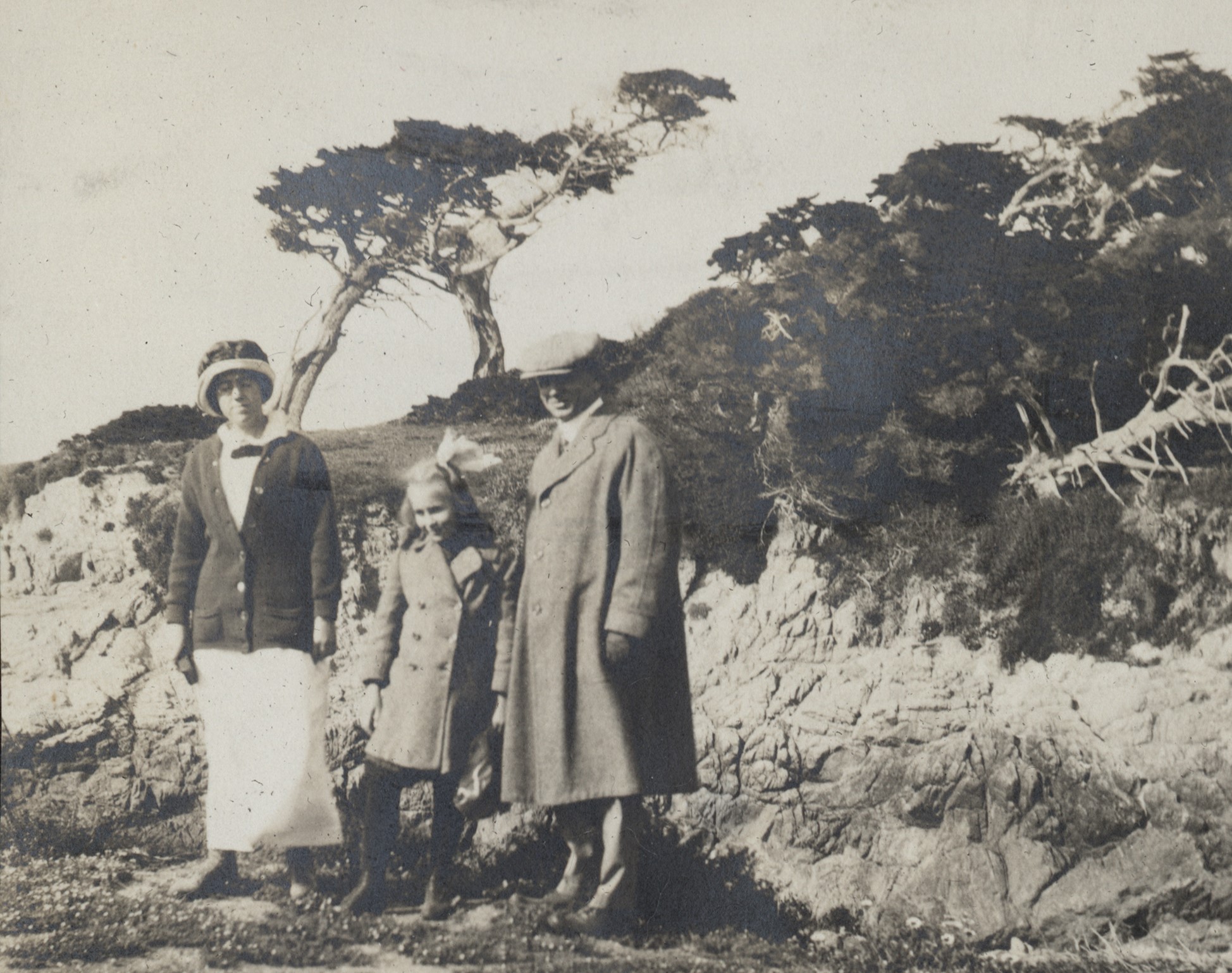 Photo of a man, a young woman, and a girl standing in front of a hillside upon which the famous cypress tree stands at the Pebble Beach golf course in California. The man wears a flat cap and a long overcoat with trousers. He's smiling, as are the girls, and he's looking at the cameraperson. The young girl stands between the other two, wearing a bow in her long hair and a coat. The young woman wears a long white skirt, a cardigan over a white blouse, and a narrow-brimmed hat with embellishments in front.