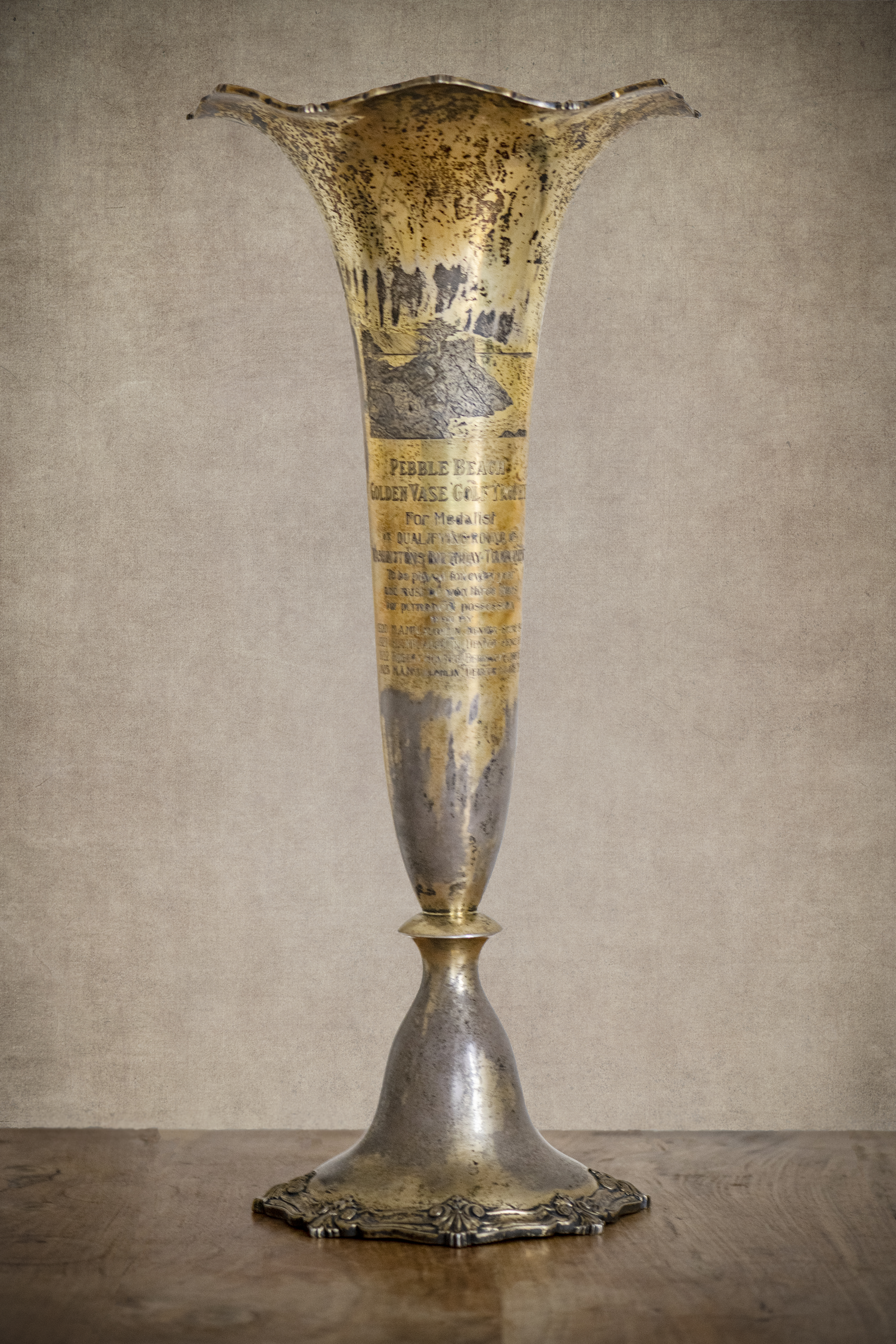 Photo of a tall slender silver-plated vase, showing some tarnish around the base. It has an image of a cypress tree on it, and is the original trophy presented to the winner of the Pebble Beach Golf Tournament, which began in 1920. The inscription shows that M.A.McLaughlin won the trophy in 1920, 1921, and 1923. A Robert Hunter won it in 1922.