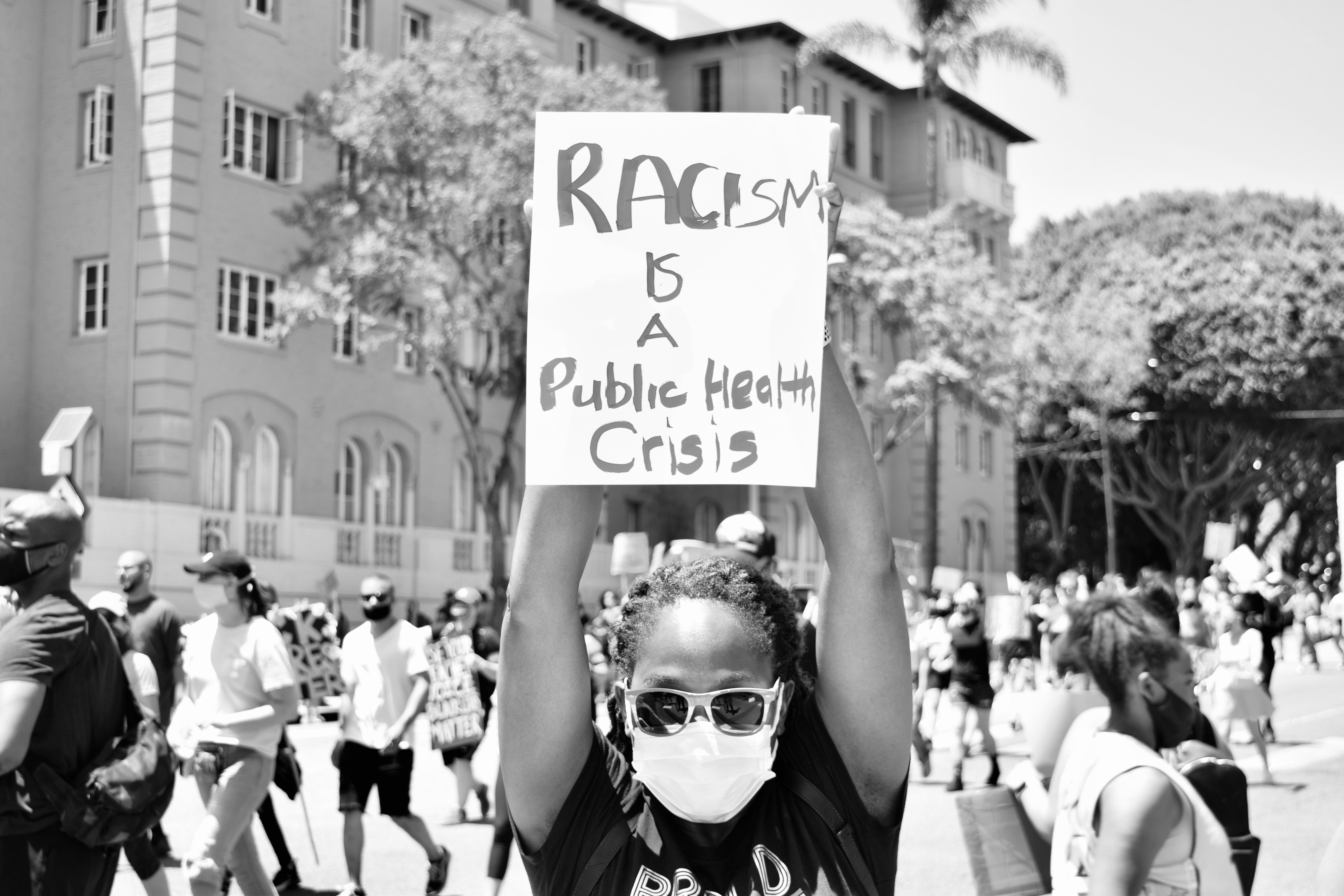 Photo of a Black Lives Matter protestor, holding up a sign that reads, "Racism is a Public Health Crisis." This person has long hair that appears to be in braids and pulled back behind their head.  They are wearing a mask due to the Covid-19 pandemic, as well as mirrored sunglasses.  They are facing the camera and holding the sign high above their head, while behind them dozens of people are marching and holding protest signs.