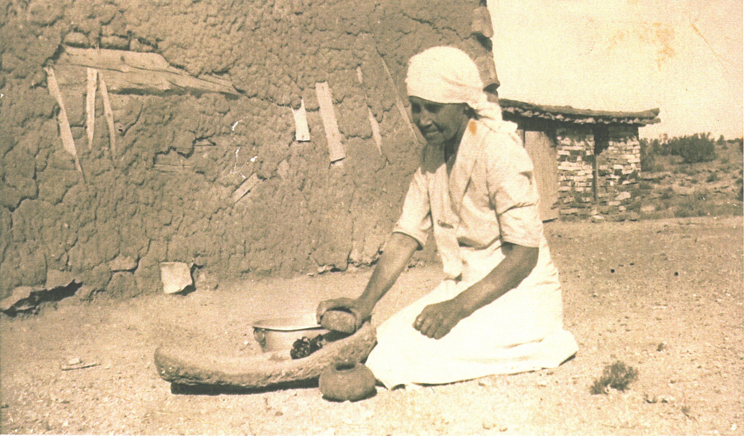 Photo of a woman sitting on the ground in front of a grinding stone, preparing to grind up a small amount of food. Next to her left of the grinding stone there sits a small pottery vessel, and to her right of the stone sits a metal pot that has a small ring handle. The woman is wearing a white skirt and blouse, and her head is wrapped in a white fabric covering that trails past her shoulders down her back. There is an adobe building behind her, and two other buildings behind her in the distance.