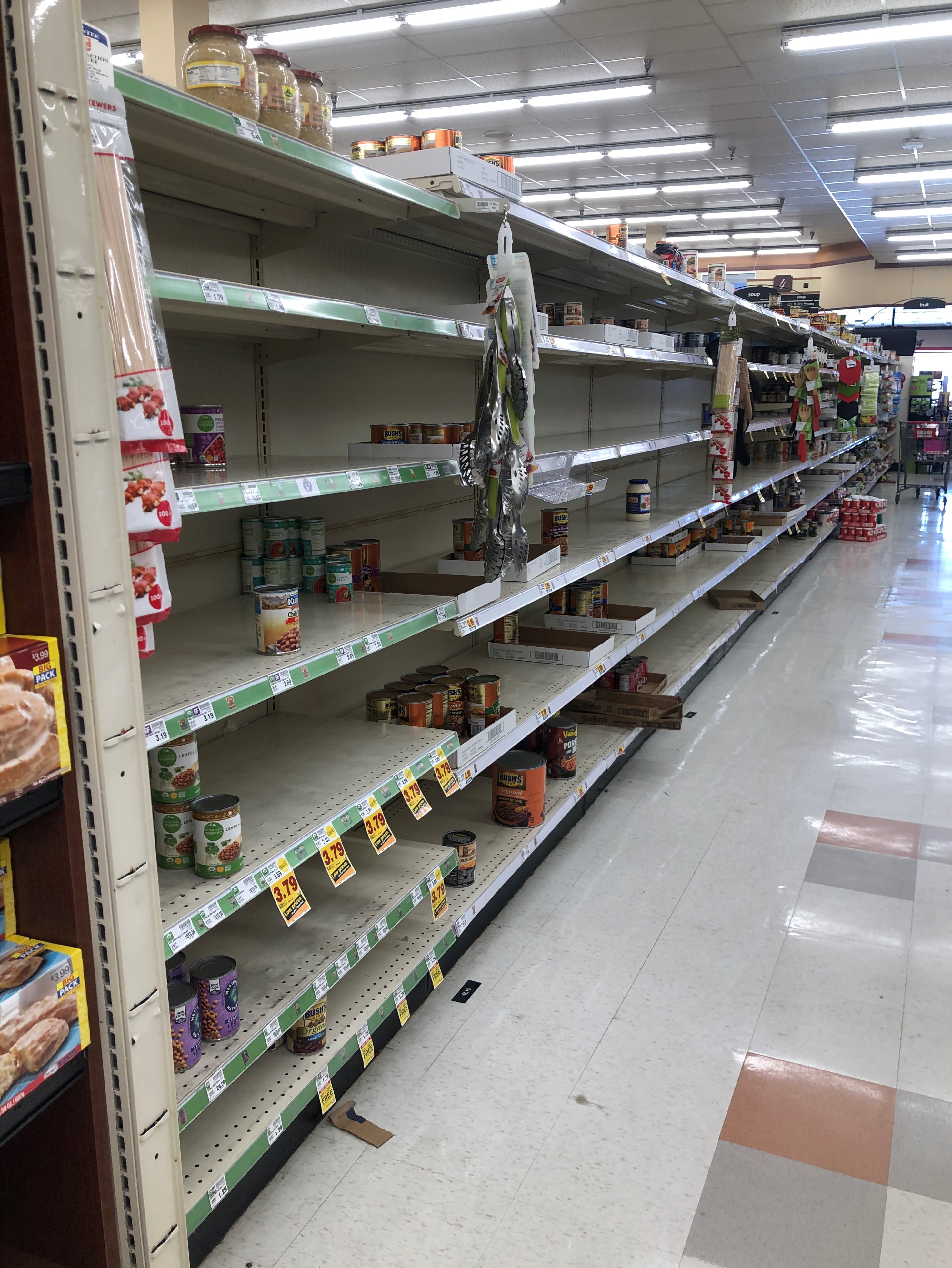 Photo of angle view of shelving down left side of canned good aisle in grocery store. Limited amount of canned beans and tomatoes are scattered throughout shelves. Shelves below "fruit" sign at opposite end of aisle appear to be more fully stocked. Shopping cart sits at far end of aisle.
