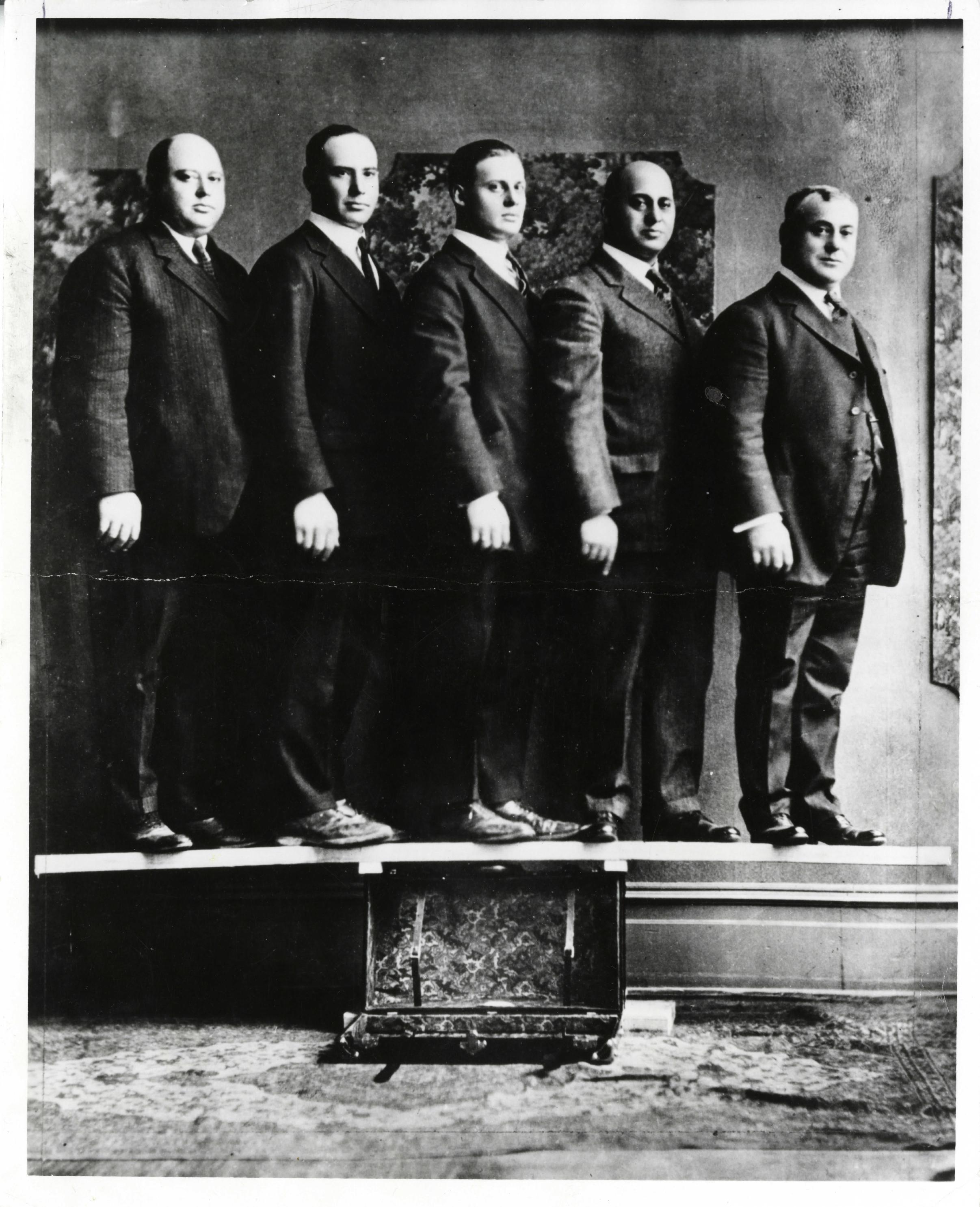 Photo of the five Shwayder brothers--Mark, Maurice, Benjamin, Jesse, and Solomon--standing on a board that is balanced on a Samsonite suitcase. This was a famous advertising image used in the early twentieth century, that the Samsonite company used to demonstrate the strength of their suitcases.