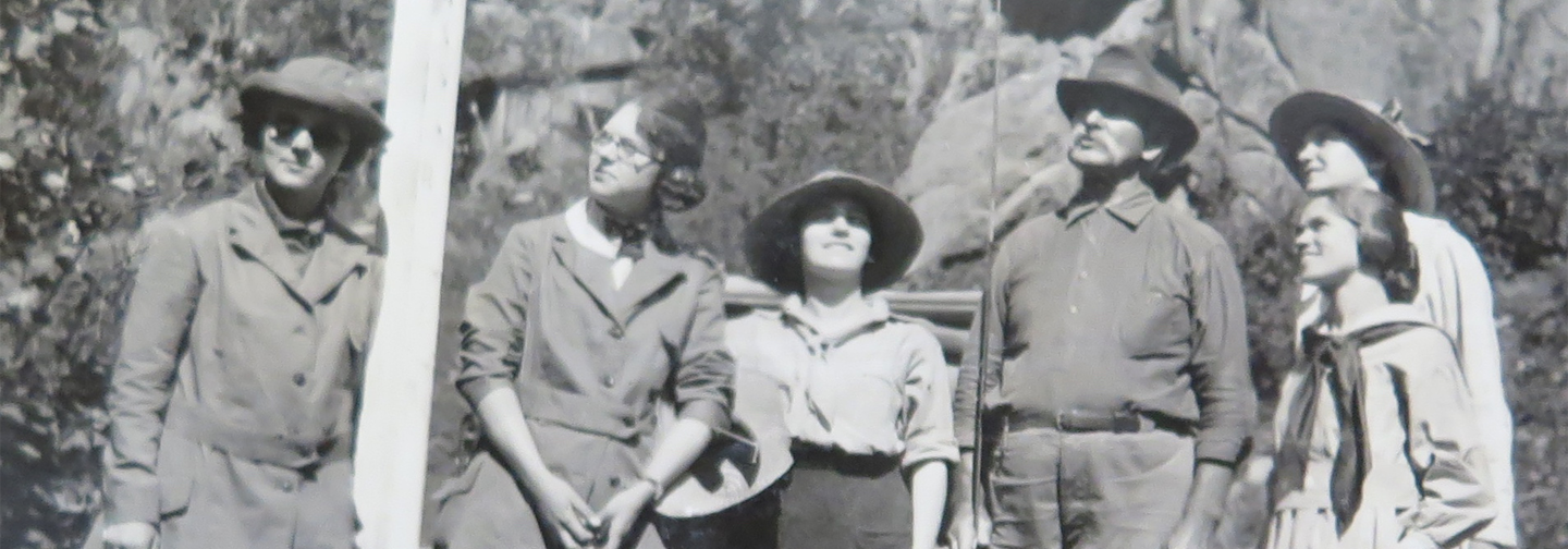 Photo of 5 women and 1 man, standing together outdoors, as if they are getting ready to go somewhere. A few of them are looking up, as if they are looking in a tree. They are wearing jackets and most of the party wears hats, and 1 woman appears to hold a camera in her hand. The man holds what looks to be a fishing pole, standing upright, in his right hand. A rocky mountain wall is visible in the background.