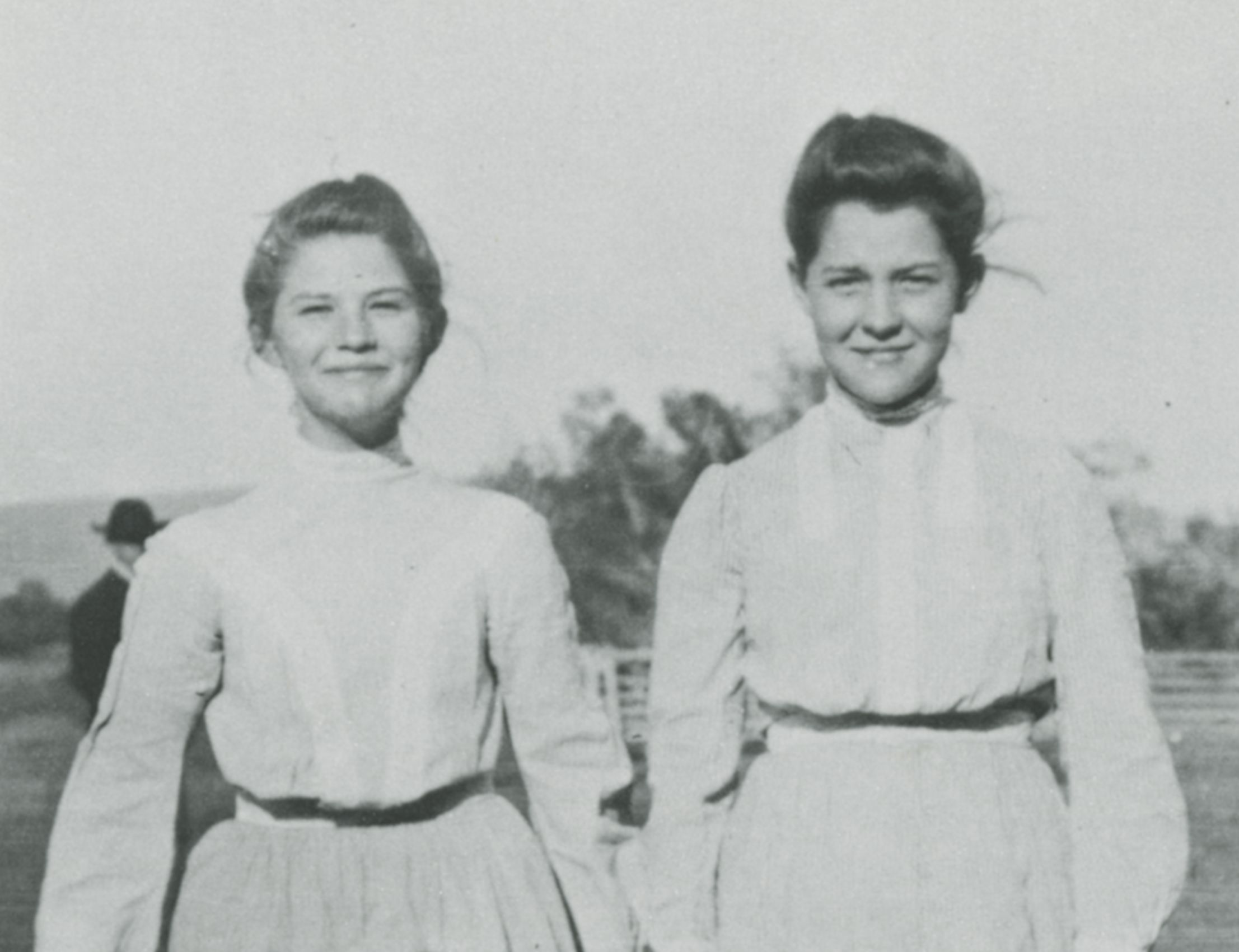 Photo of two young women standing side by side, outdoors. They are both wearing light colored, high neckline, long sleeved blouses with skirts, and they both wear their hair pulled up into buns on their heads. They are smiling at the camera.