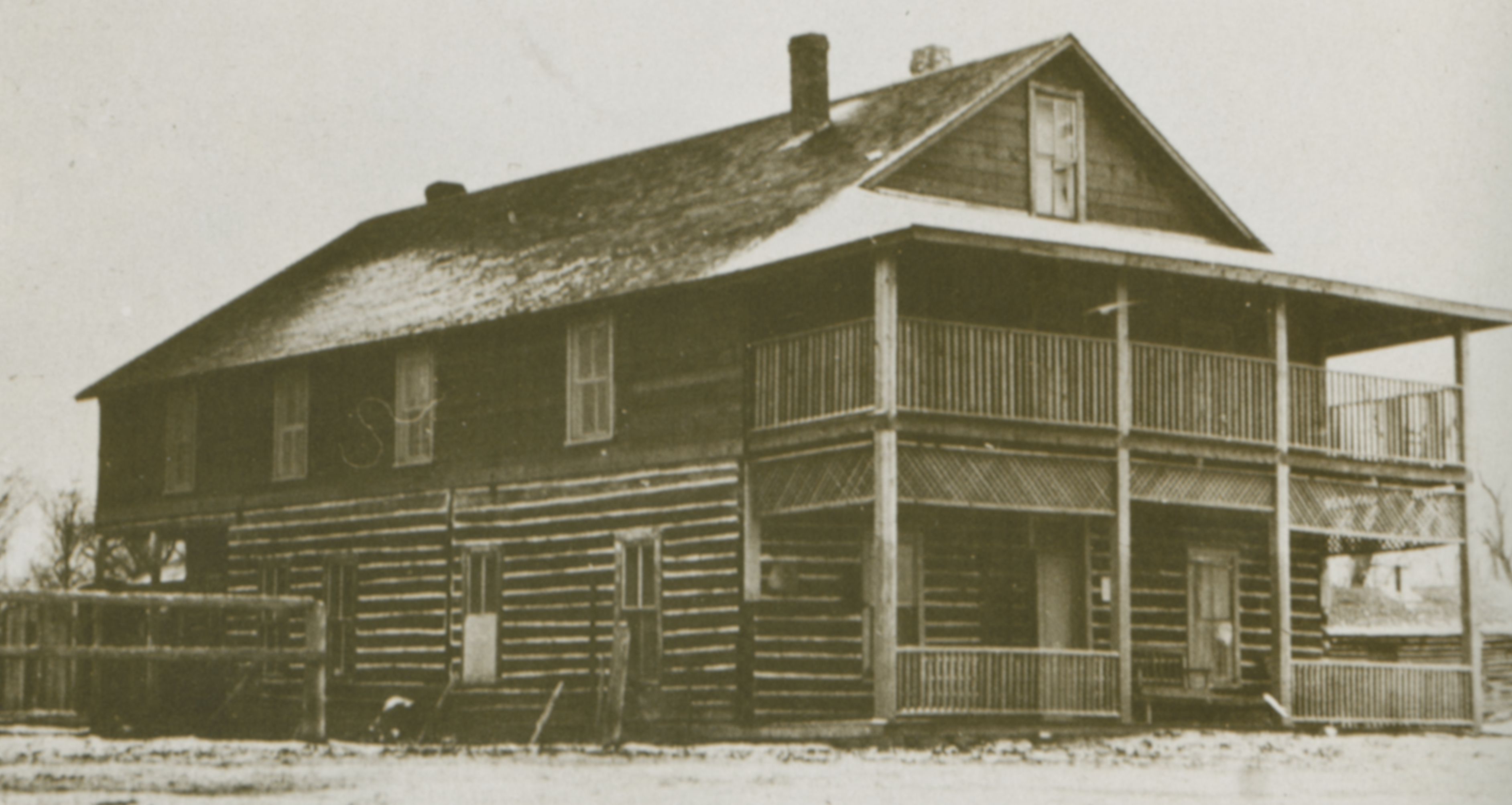 Photo of a two-story log home, with covered porches on the short end of this rectangular building. It has a simple pitched roof from which the chimney stands. The log structure has several small window on both floors  and on the two sides of the home that are visible. The yard has fencing, separating land to the rear of the house from the front yard area. A dusting of snow decorates portions of the rooftop.