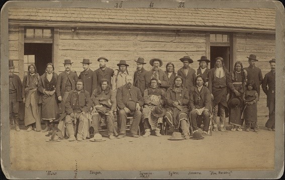 Image of a historic cabinet card showing a large group of people in front of a long cabin. The group includes John Taylor, Black Civil War veteran. He stands in the back row, alongside others who are gathered and posing for the photo, including Ute Chiefs Ignacio and Severo, and others in the diverse group of  men and women.
