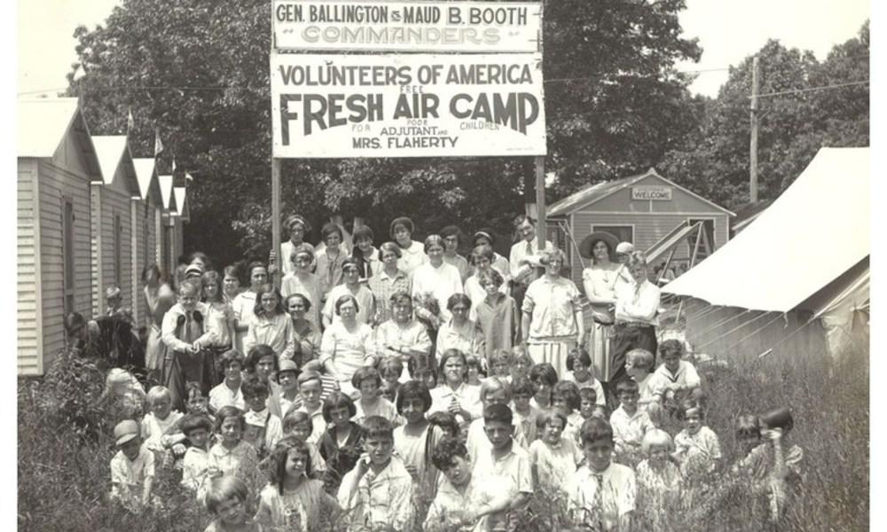 Photo of a large group of adults and children, posed in front of and under a large overhead sign that reads, "Gen. Ballington & Maud B. Booth, Commanders - Volunteers of America Free Fresh Air Camp for Poor Children." The campers are around ten years in age. A tent and several small cabins are adjacent to the group standing in the field.