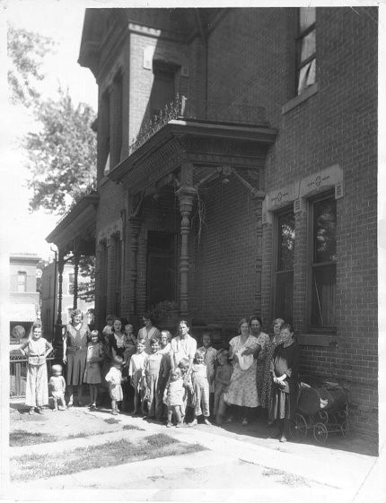 Photo of a two-story brick building, likely built during the late-nineteenth or early-twentieth century. Ornate ironwork and wooden accents on the building are visible. There is a group of about 20 women and children standing in front of the building and posing for this photo.