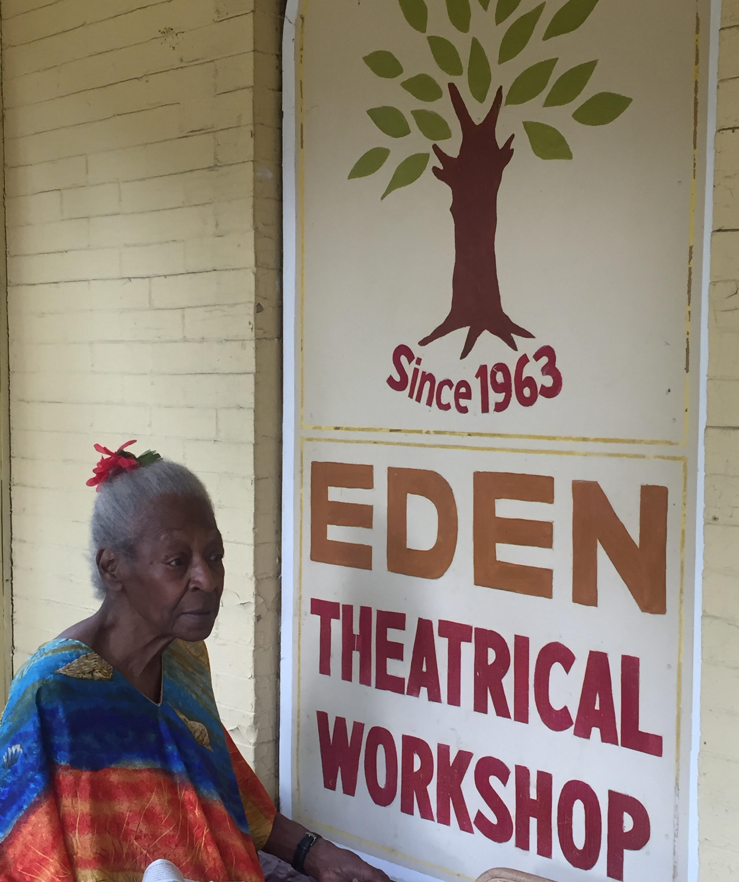 Photo of Lucy Walker, an older Black woman, standing in front of the sign on the front of her business. The brick building is painted yellow & the sign has a tree at the top of it. Below the tree trunk says "Since 1963." Below that, large yellow block letters say "EDEN," followed by "Theatrical Workshop" red block letters. Lucy is looking off to the right. Her gray hair is in a bun atop her head, tied with a red ribbon. She wears a blouse of bright blues, reds, and oranges.