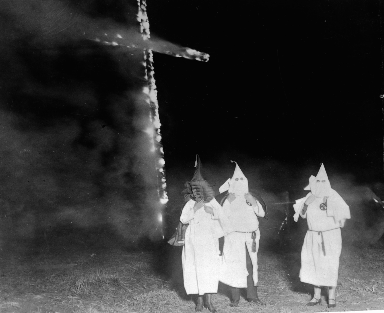 Three members of the KKK stand near a burning cross at night