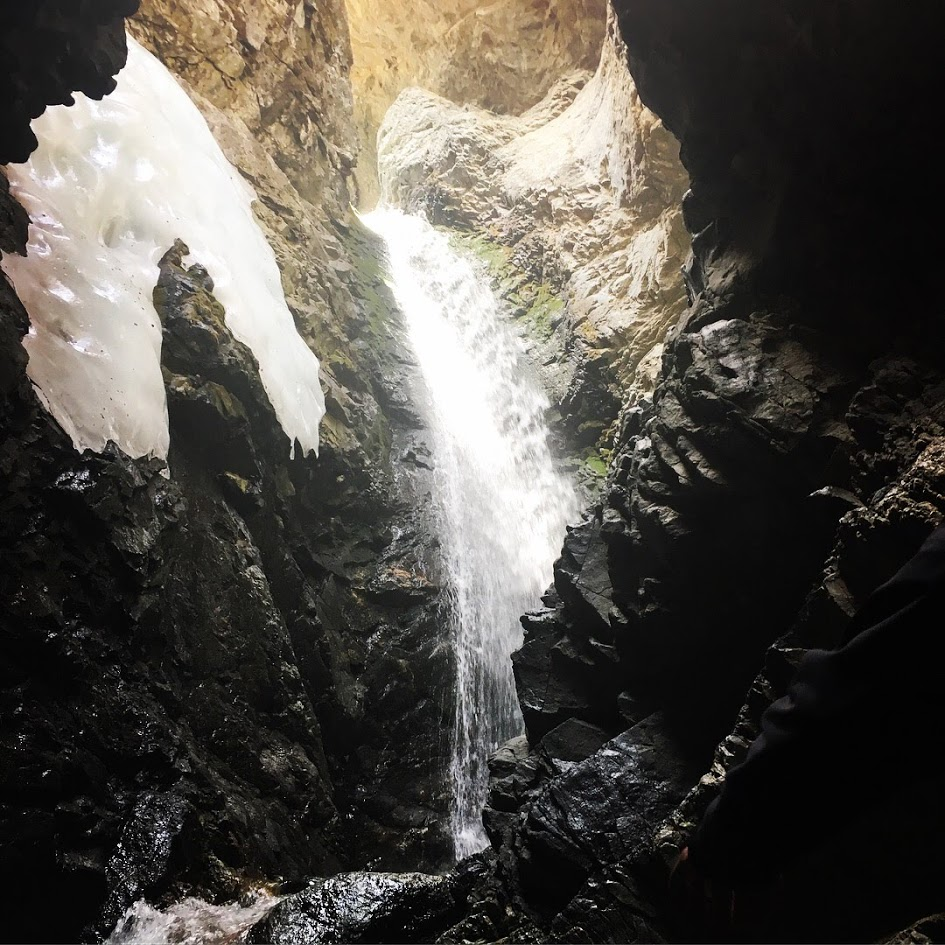 Photo of a waterfall, spilling into a rocky underground mountain crevace. The sun is shining above, where the rocks are a lighter color and the water comes from. A patch of smooth white ice is attached to the left side of the cavernous wall.