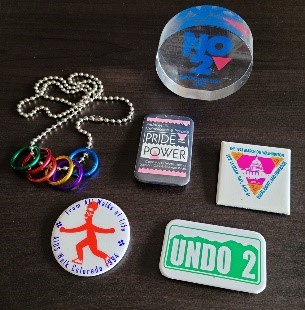 Photo of History Colorado collection items pertaining to the historic Amendment 2 Repeal. Items are: a "No on 2" clear round paperweight, pictured standing up on its side; a rectangular button with a design that looks like the iconic Colorado mountain license plates, and says "UNDO 2"; a silver metal chain on which there are 6 thin metal rings, each a different color of the rainbow, and 3 other buttons, for AIDS Walk 1994, PRIDE Power, and another (not legible).