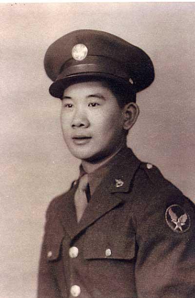 Photo of an Asian man in a miliary uniform, posing for his portrait. He wears a military hat with a single large metal medallion in the center above the brim. His lapeled jacket has two pockets with flaps that are fastened by metal buttons, and is buttoned up with metal buttons. There is a round patch on his left upper sleeve, that shows a white star with a circle in it, and two wings on the star. He wears a dark colored dress shirt and a lighter colored necktie. There is a lapel pin on his left collar.