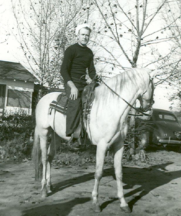 Photo of a man in black trousers and a black shirt with what looks like a white sailor-style hat, sitting atop a white horse. The horse is standing under some trees which are in front of a small house. A car is parked nearby in this vintage black and white photo.