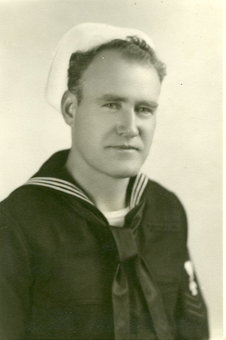 Photo of a man in his Navy uniform, posing for a military portrait. His dark shirt has a sailor collar edged by 3 white stripes, with a tie in front. He is wearing a white sailor's cap and is looking at the camera.