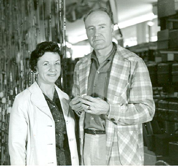 Photo of a woman with dark hair, on the left, standing next to a man with light hair on the right. The are looking at the camera. They are wearing shirts with light jackets over them. The man looks like he is holding something small in his hands.