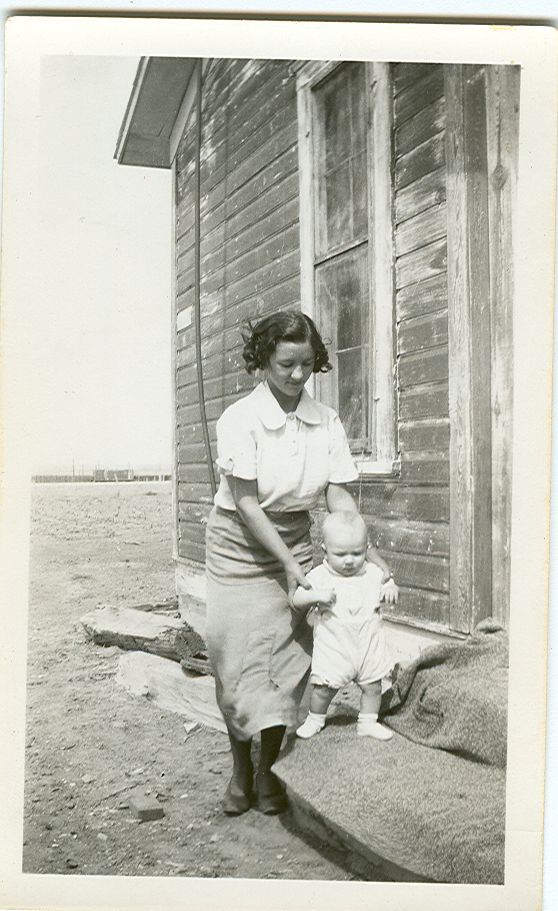 Photo of a young woman supporting the arms and shoulders of an infant who is standing on a stairstep and trying to walk. The step is part of a wooden cabin-like structure with wooden siding and a window on the wall behind the woman. The ground around the structure is flat dirt in this vintage black and white photo.