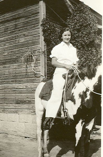 Photo of a dark-haired woman sitting atop a horse with the reins in her hands. The horse is standing in front of the corner of a small building with wooden siding and vines growing up one side of the structure. She is looking to the right of the image, and smiling. She is wearing a light-colored, short-sleeved blouse and light-colored trousers in this historic image.