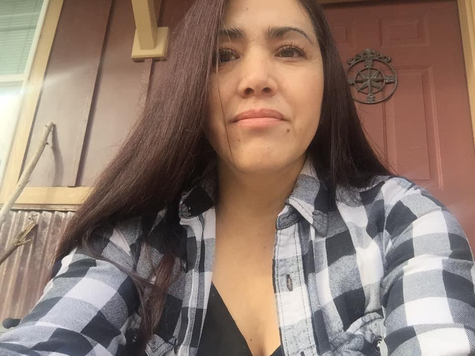 Sandra Lopez sits in front of a door. She wears a black and white plaid button down open with a black shirt underneath. 