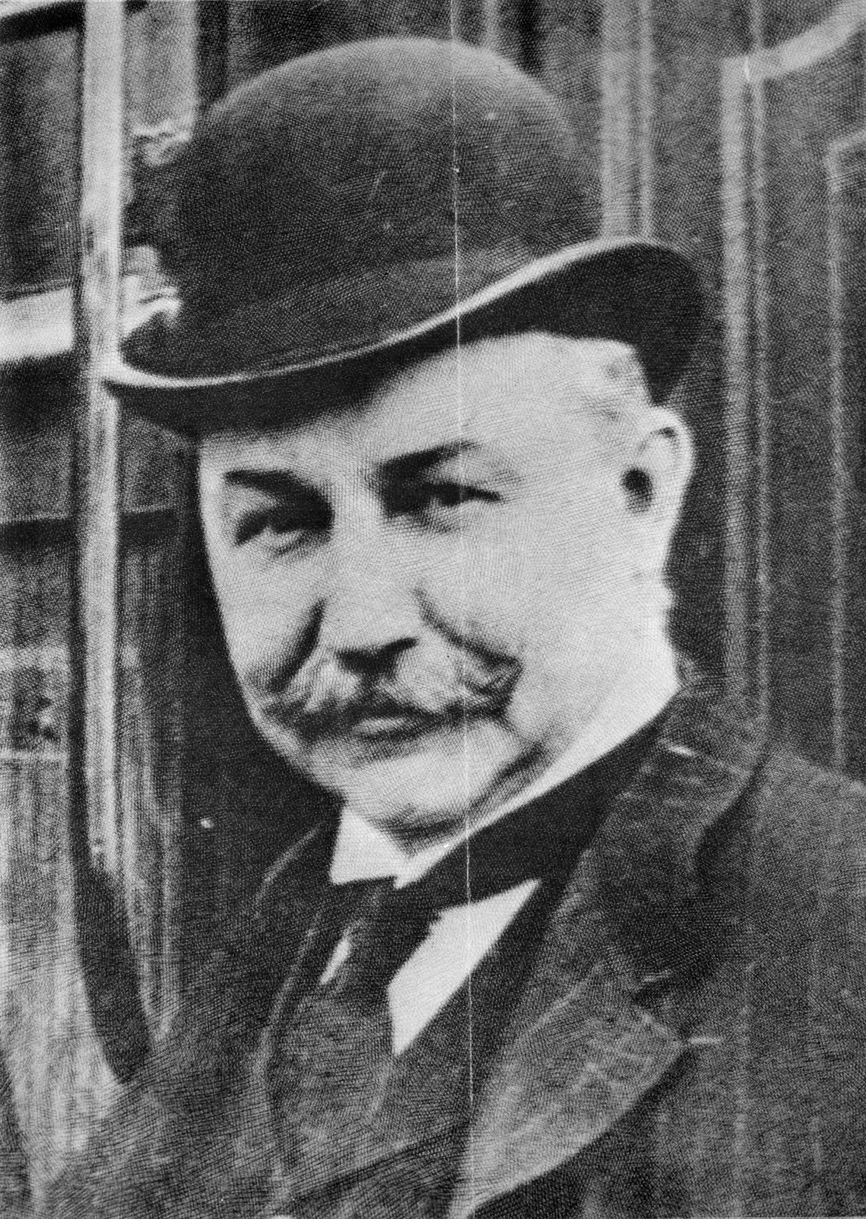 A black and white photo of a man in a suit. He wears a mustache and a bowler hat.