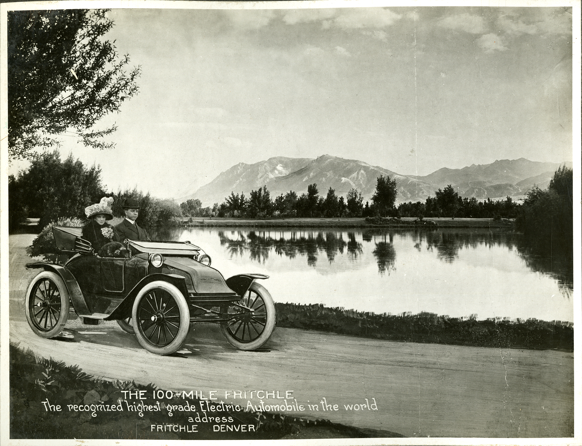 A man and a woman in turn-of-the-century dress ride in an early open-top car. The car is driving along a gravel path next to a small body of water. Mountains are on the horizon. A caption reads "The 100-mile Fritchle: The recognized highest-grade electric automobile in the world. Address: Fritchle, Denver."