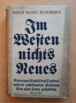 Photograph of a book with a worn white cover. The words on the cover are all in German, and in the center in large black letters the cover of the book reads, "Im Westen nichts Neues" which translates to "All Quiet on the Western Front."  Red block letters across the top of the book spell out the author's name, Erich Maria Remarque. 