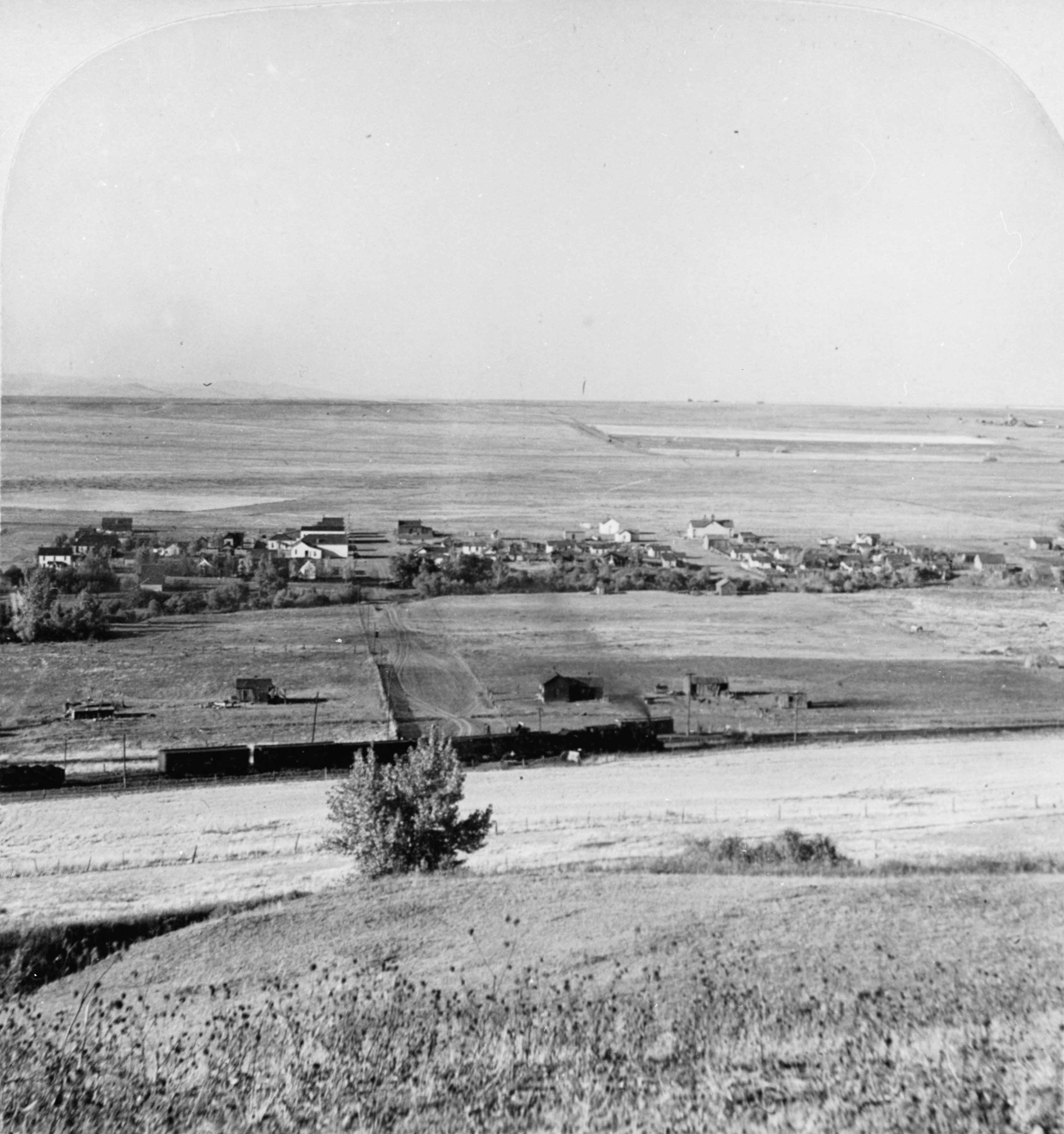 A historic photo of a small town with the prairie stretching out behind it. The railroad runs parallel to the town.