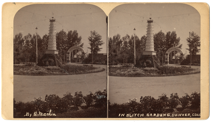 Photo on a vintage stereoscope card. The image is labeled by hand at the bottom with the words "By A. Martin" and "In Elitch Gardens, Denver, Colo." The photo shows a tall white round tower, like a lighthouse, with a spiral row of light bulbs ascending the tower. It sits on a square pedistal that is in the center of a flowerbed. Four strings of lights attach to the top of the tower. An archway in the background says "Elitch Gardens."