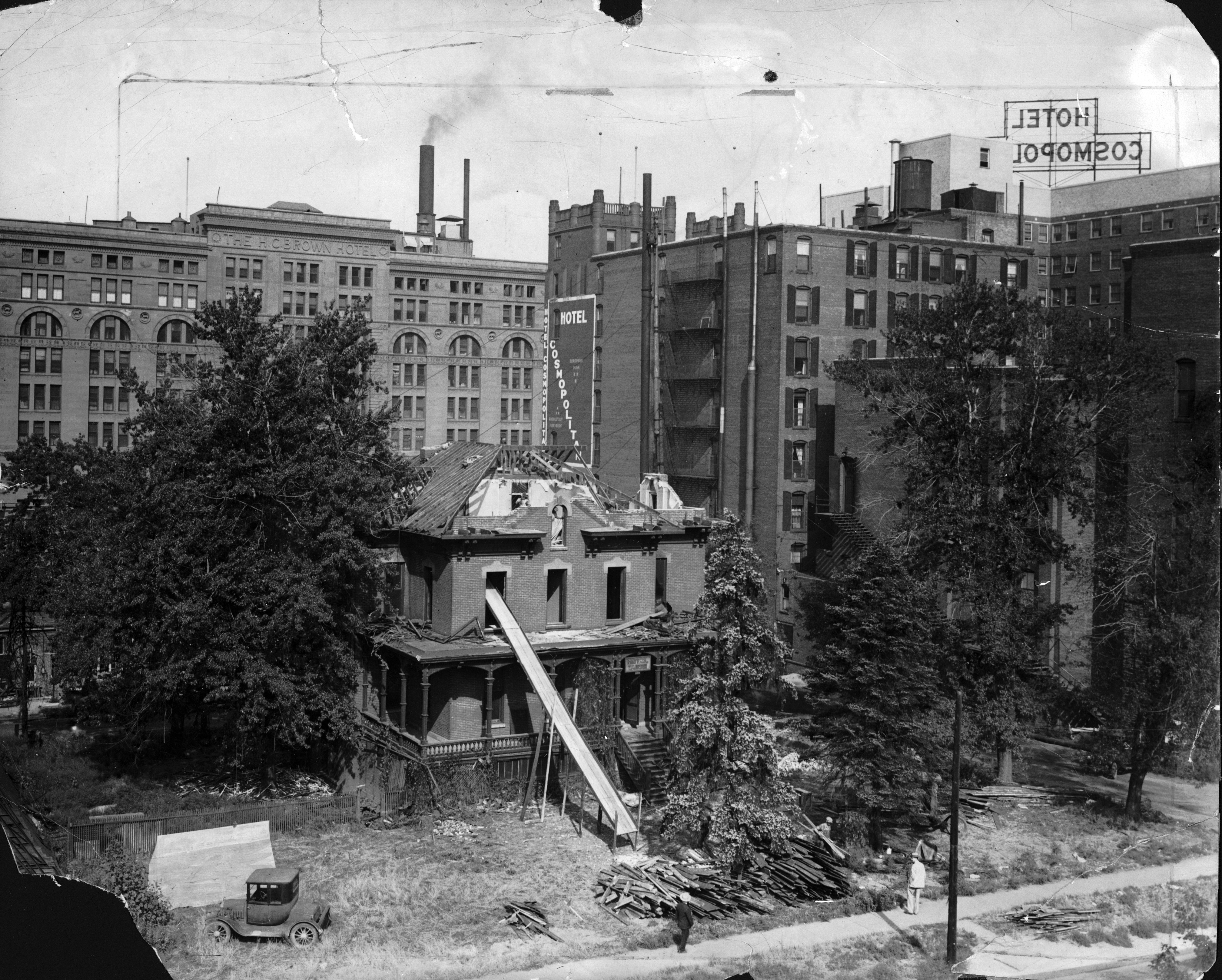 Photo of a Victorian home being demolished, with multi-story buildings towering over it. There is a chute coming from the front upstairs window of the house, down to the front lawn. The roof is being torn down and large parts of it are missing, exposing wooden beams. All of the windows are gone. Tall trees stand near the house, and an vintage car is in the corner of the front yard. Atop a tall building in the distance are tall letters that say "Hotel Cosmopolitan."