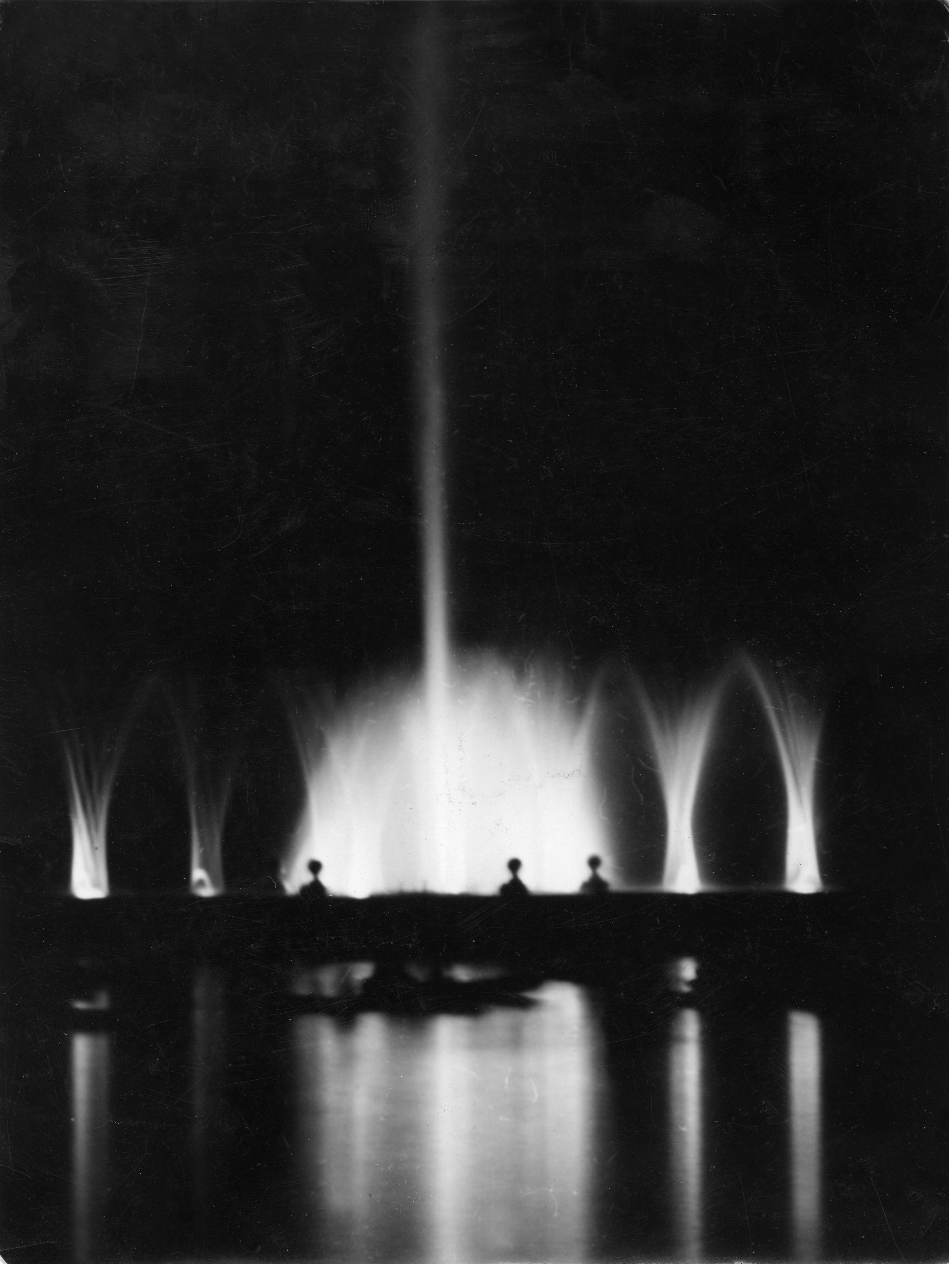 Photo of fountains at Denver's City Park, illuminated in the nighttime darkness. There are two smaller sprays up into the air on each side of a larger cluster of water sprays, with one tall burst of water in the center. The water glows white, and the reflection of the illuminated fountain sprays are visible in the perfectly still water of the lake in the foreground.