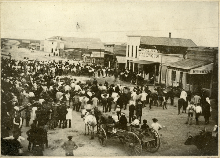 Photo of a crowd gathered in front of one-story Old West-style buildings with signs that read "Wholesale & Resale: Clothing Store" and "Confectionary - Ice Cream." Along the dirt street, a crowd has gathered in a circle below a tiny figure high above the crowd. The figure balances along a tightrope stretched between what is likely to be two buildings, although the photo does not show what the rope is secured to. There is a horse and cart in the front and oxen to the right corner of the foreground.