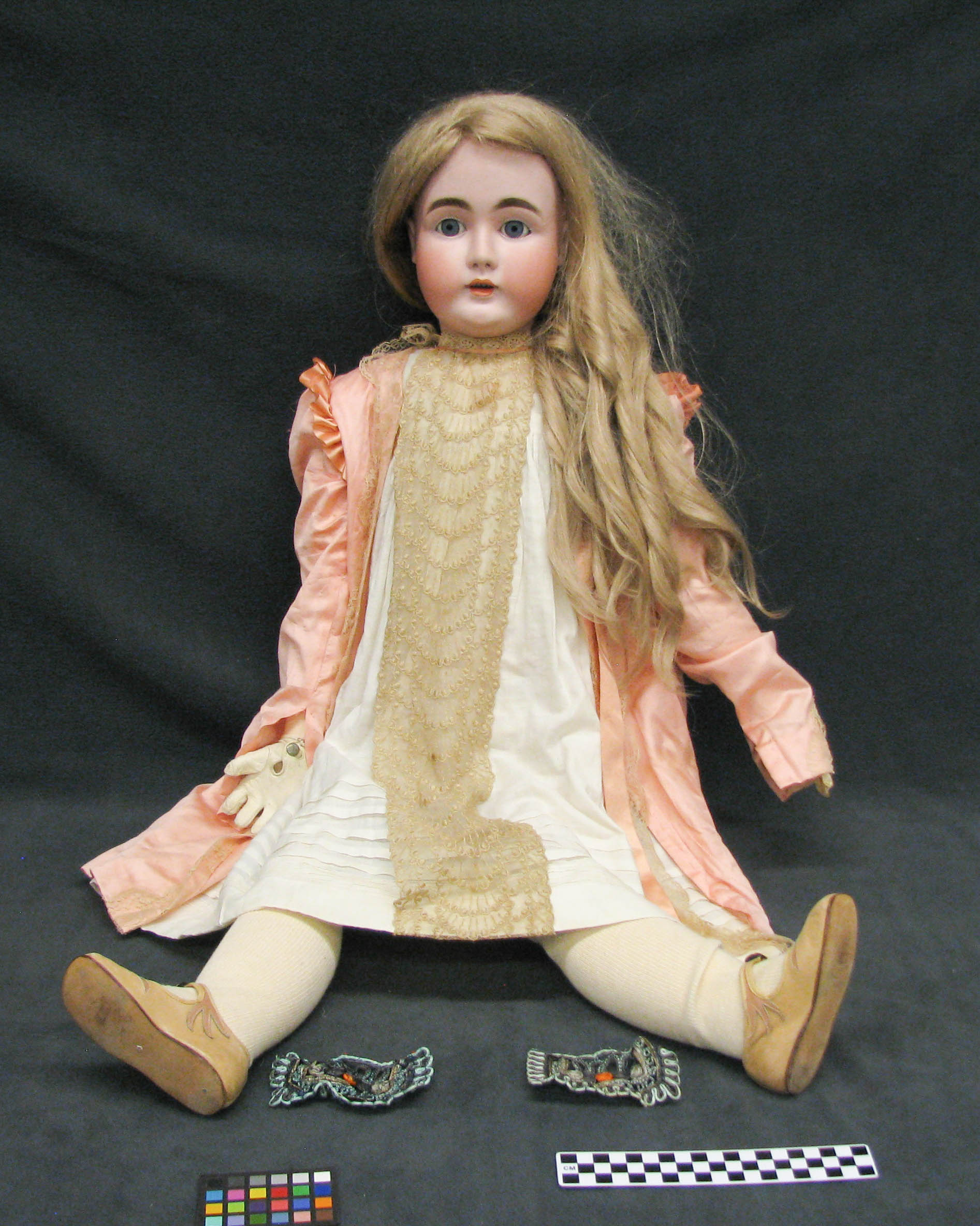 Photo of a porcelain doll, sitting upright against a black draped background. She is wearing a pink dress and shoes with white gloves and stockings. She has long blonde hair that falls in ringlets over her shoulders. She has blue eyes, pink full cheeks, and painted brown eyebrows. She is placed next to color swatches and measuring strips to document her size and colors for the purpose of the museum collection.