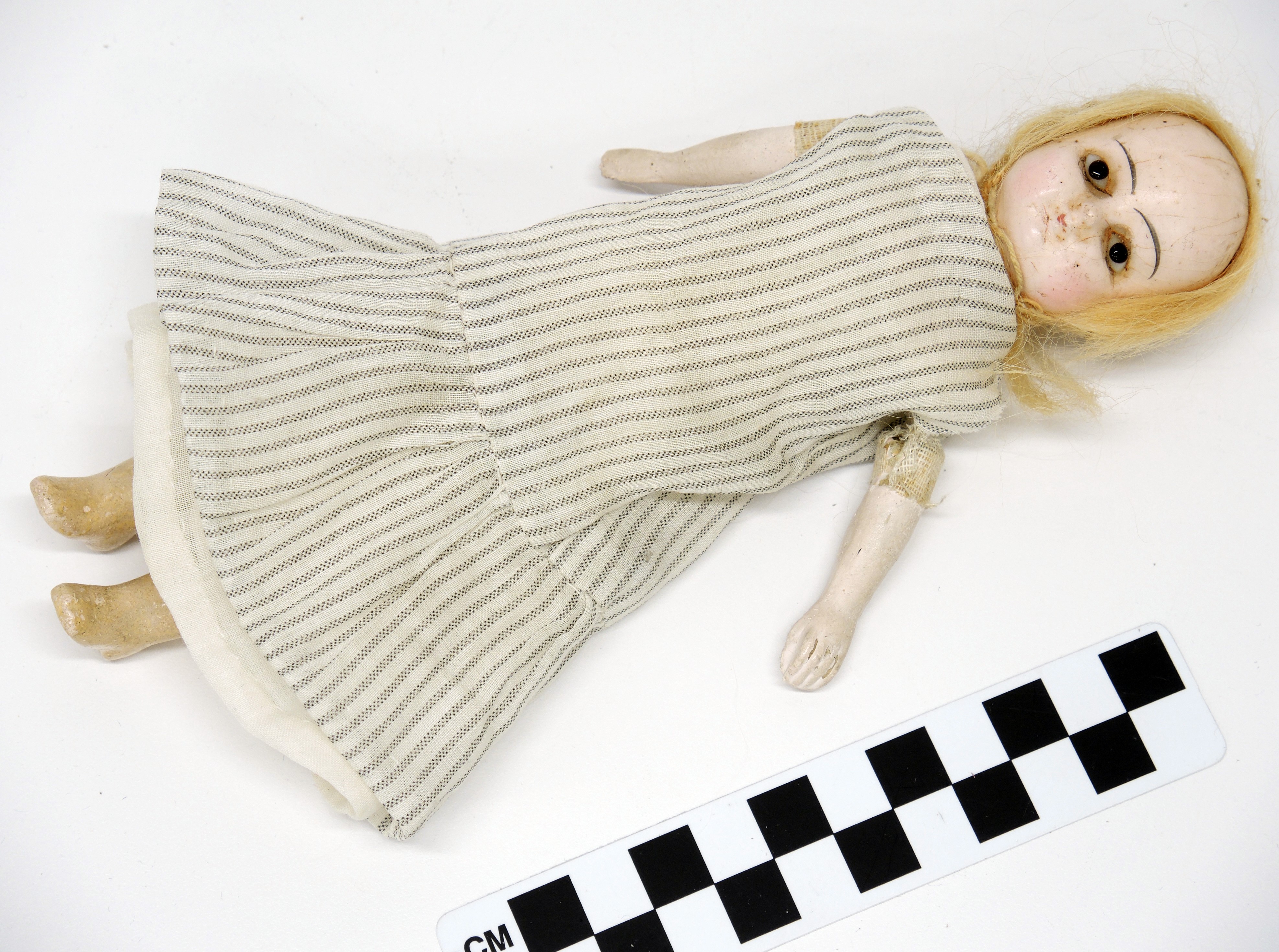 Photo of a doll from the museum collection, lying on a plain flat white background and next to a centimeter measurement strip. The doll approximately 15-18 cm long, and has thin straight shoulder-length blonde hair. Its facial markings are simple, with painted eyebrows and no eyelashes. The doll is dressed in a thinly striped gray and white dress, simply styled and made of cotton.