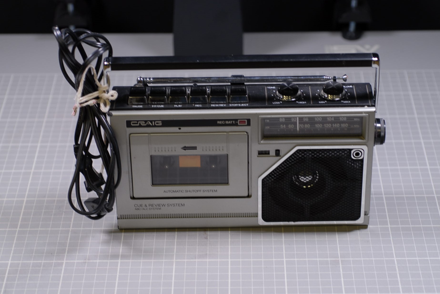 Photo of a modest-sized portable cassette tape player, photographed on a gray gridded mat to indicate the size. The cord is bundled, tied together with string to the handle. There is a cassette tape player on the left, and a window indicating radio bands, along with a silver speaker encased in a black mesh cover, on the right side of the player. Along the top of the device are buttons to operate the tape player, and two dials to control the sound. An antenna is lying flat against the top of the device.