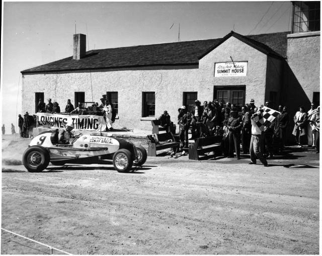 Photo of an open-wheeled race car, number 9, reaching the Summit House at the top of Pikes Peak. Spectators stand in front of the Summit House to watch the finish as someone waves the checkered flag marking the end of the driver's run in the competition. 