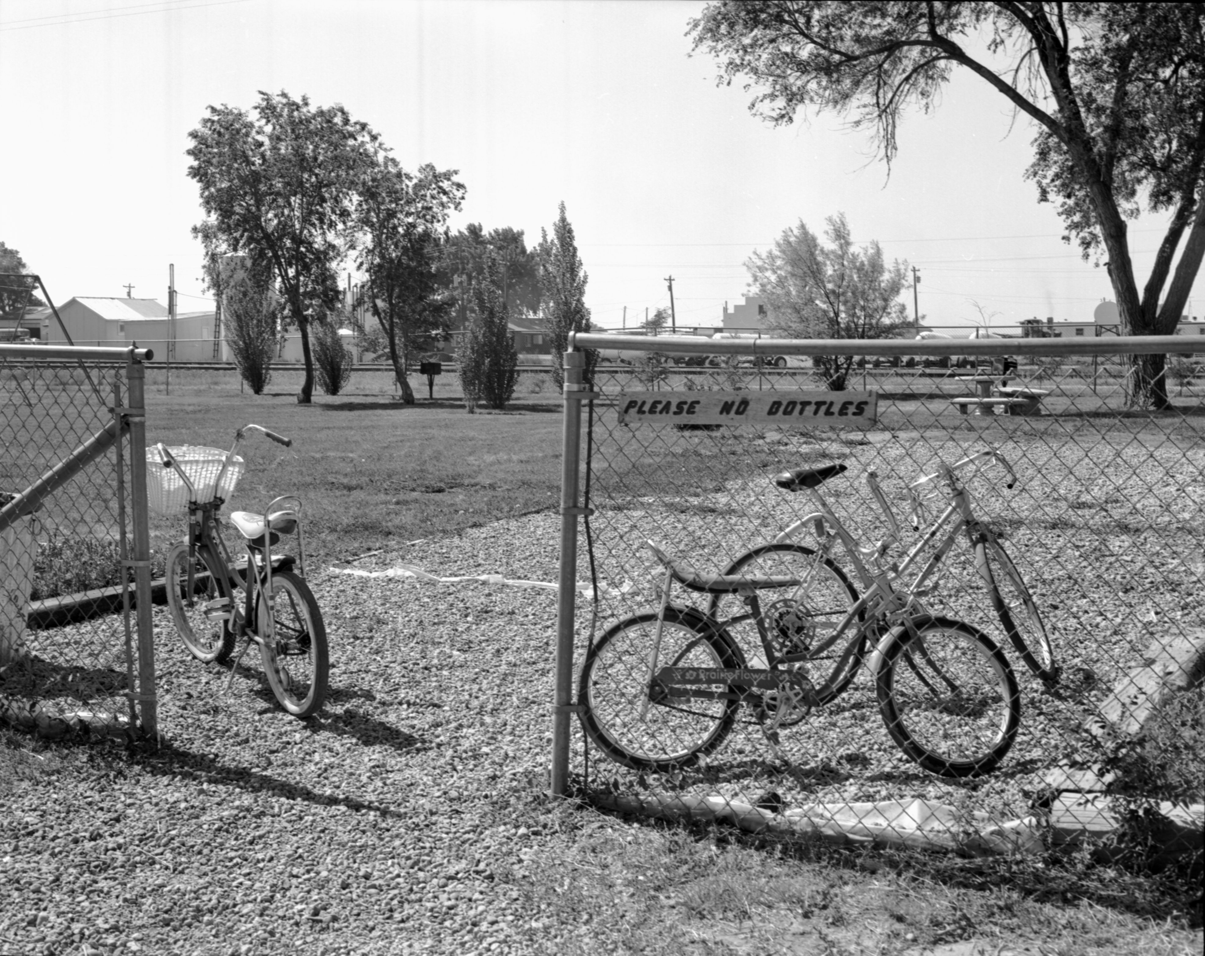 Photo of 3 kids' bicycles parked just inside of the chain link fence surrounding a municipal park. The sign next to the open gateway says "Please no bottles." The park grounds are of grass, with a few trees on the opposite side, and a round picnic table. The skies are clear and there is no one in the park on this day.