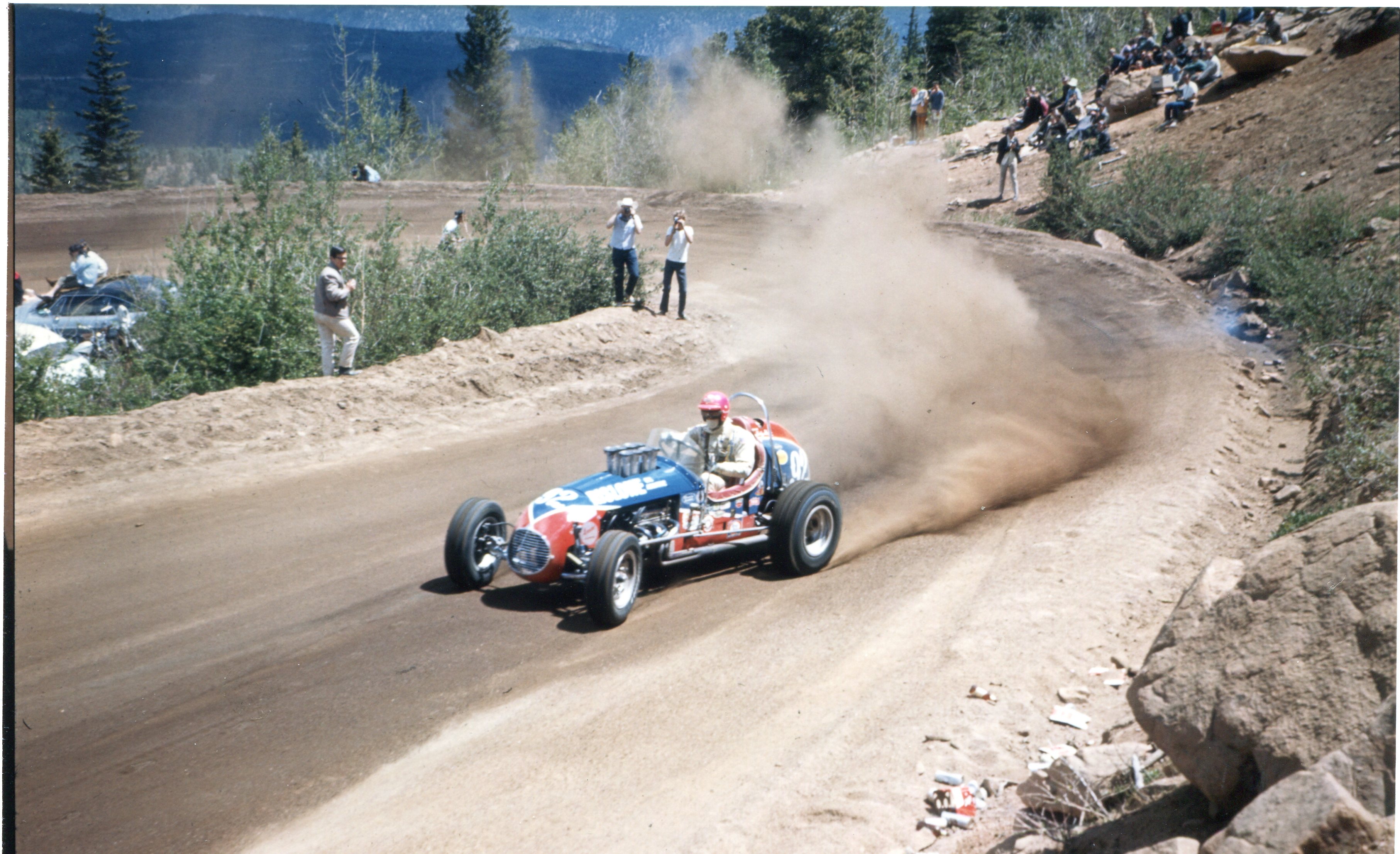 Photo of an open-wheeled race car, speeding past a hairpin turn on a dirt race track, a modest cloud of dirt kicked up behind the car. Spectators stand on both sides of the track and up the rocky hillside. The car itself is painted in red, white, and blue, although the markings are difficult to read. The driver has a white suit and a red helmet.