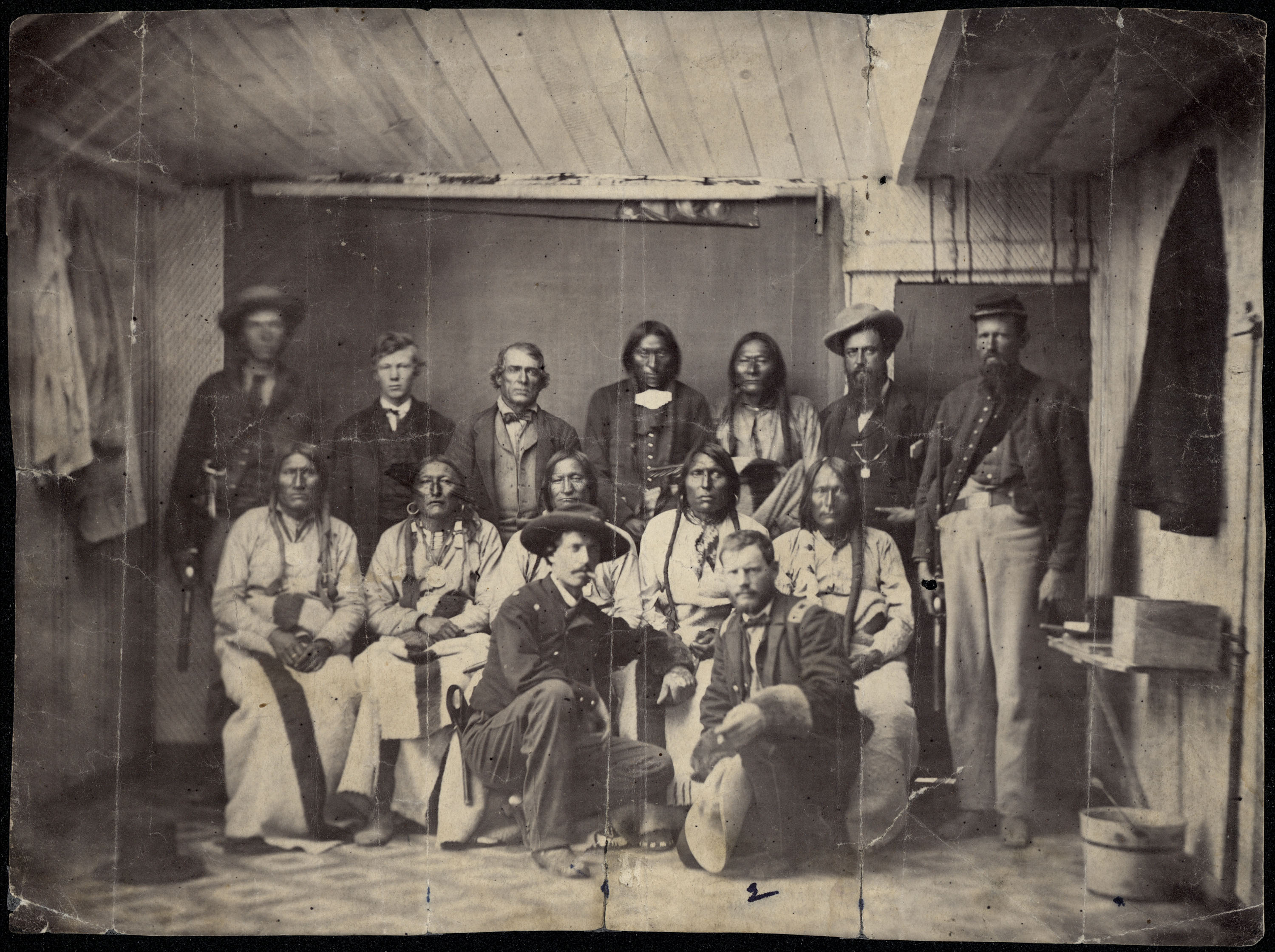 Photo of a group of about 15 men gathered in a small room for a portrait. In the foreground, two soldiers kneel in front, facing the camera. Five Indigenous people are seated in the row behind the two men. Behind them stand seven men, some of whom are Indigenous, and others who are soldiers.