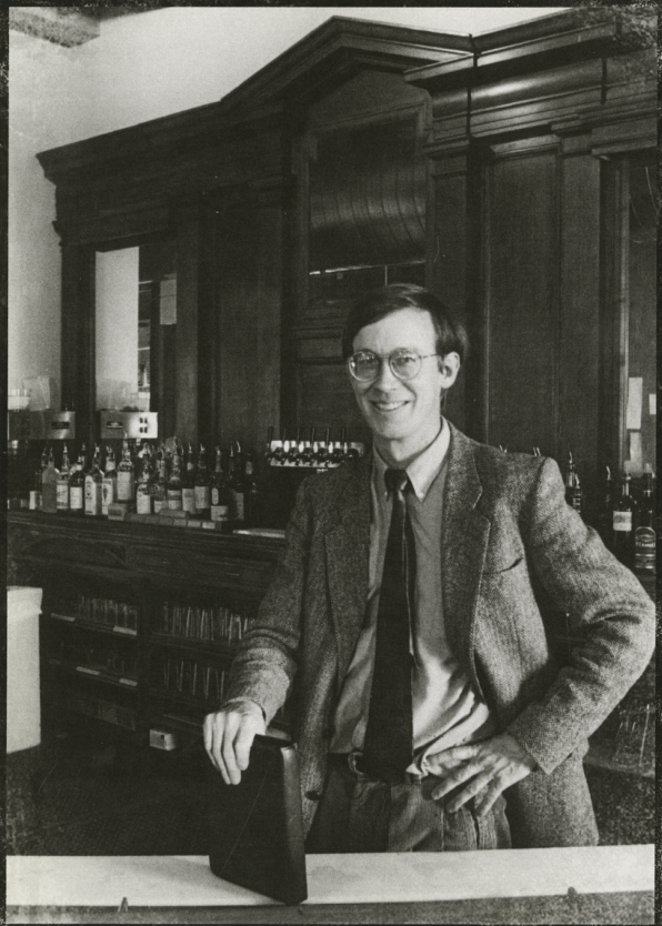A young John Hickenlooper in a suit poses at a bar with bottles of beer behind him.