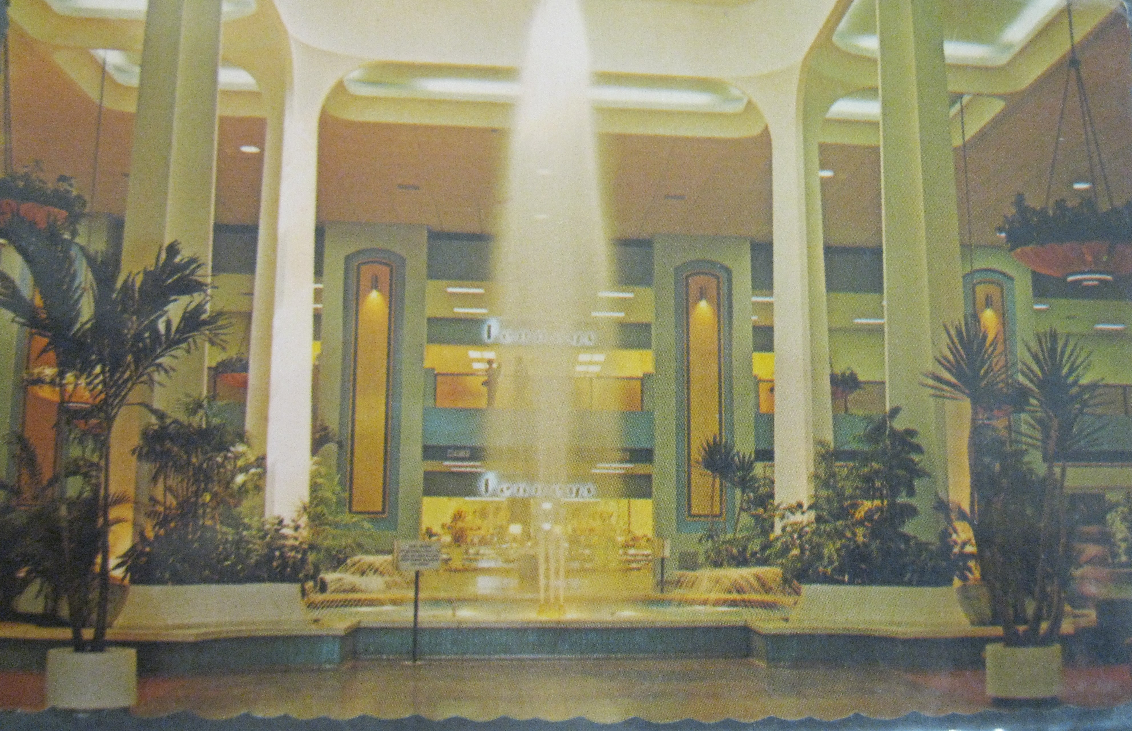 Photo of one of the giant fountains inside of the Cinderella City shopping mall. The fountain is flanked on each side by a large white structural column that reaches a very high ceiling that looks to have large sky lights directly above the fountain area. The fountain area is surrounded by large green leafed plants, and there are no people around at the time the photo is being taken.
