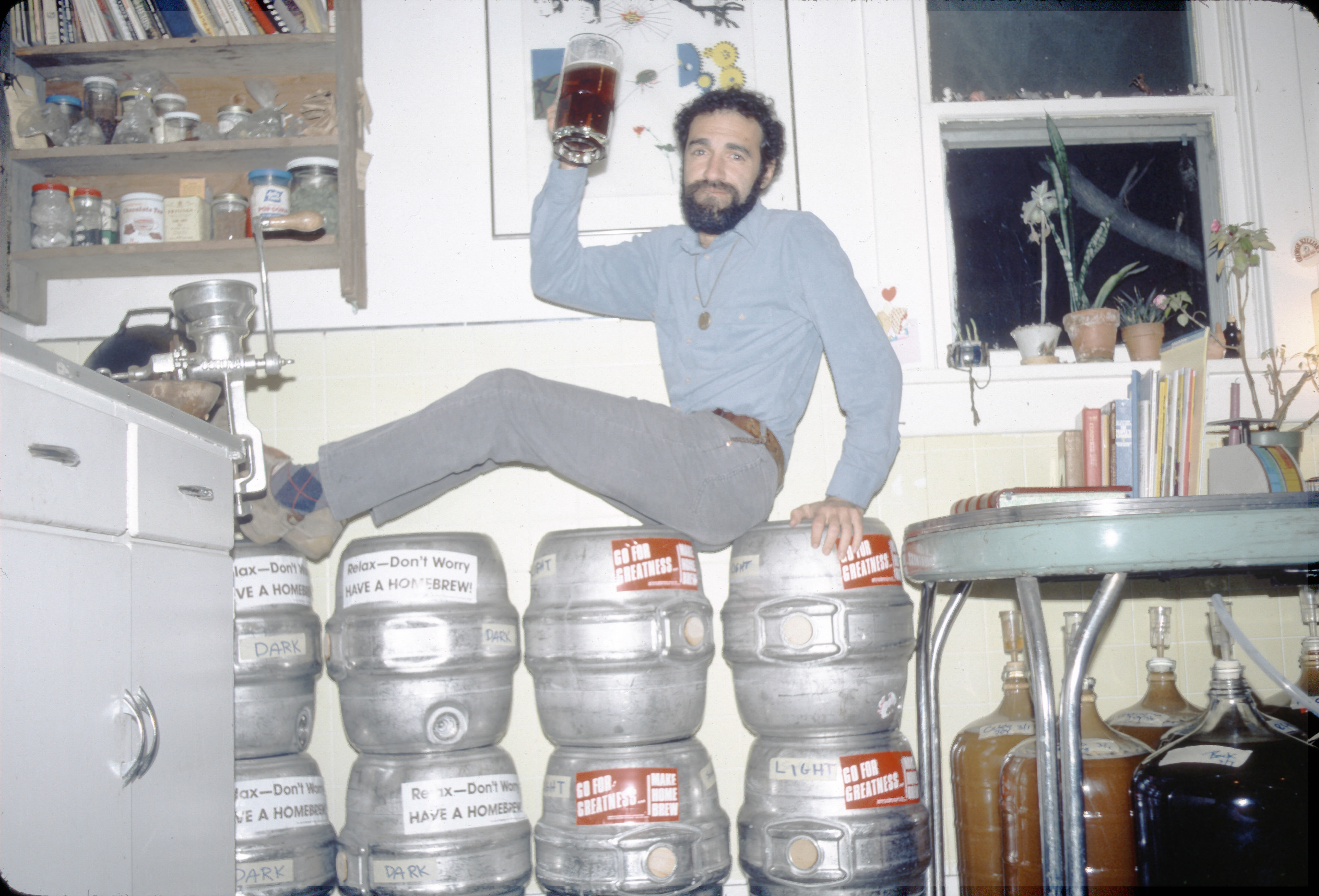 Charlie Papazian posing on a group of kegs. He holds a mug of beer, and jugs of brewing beer are visible underneath a nearby table.