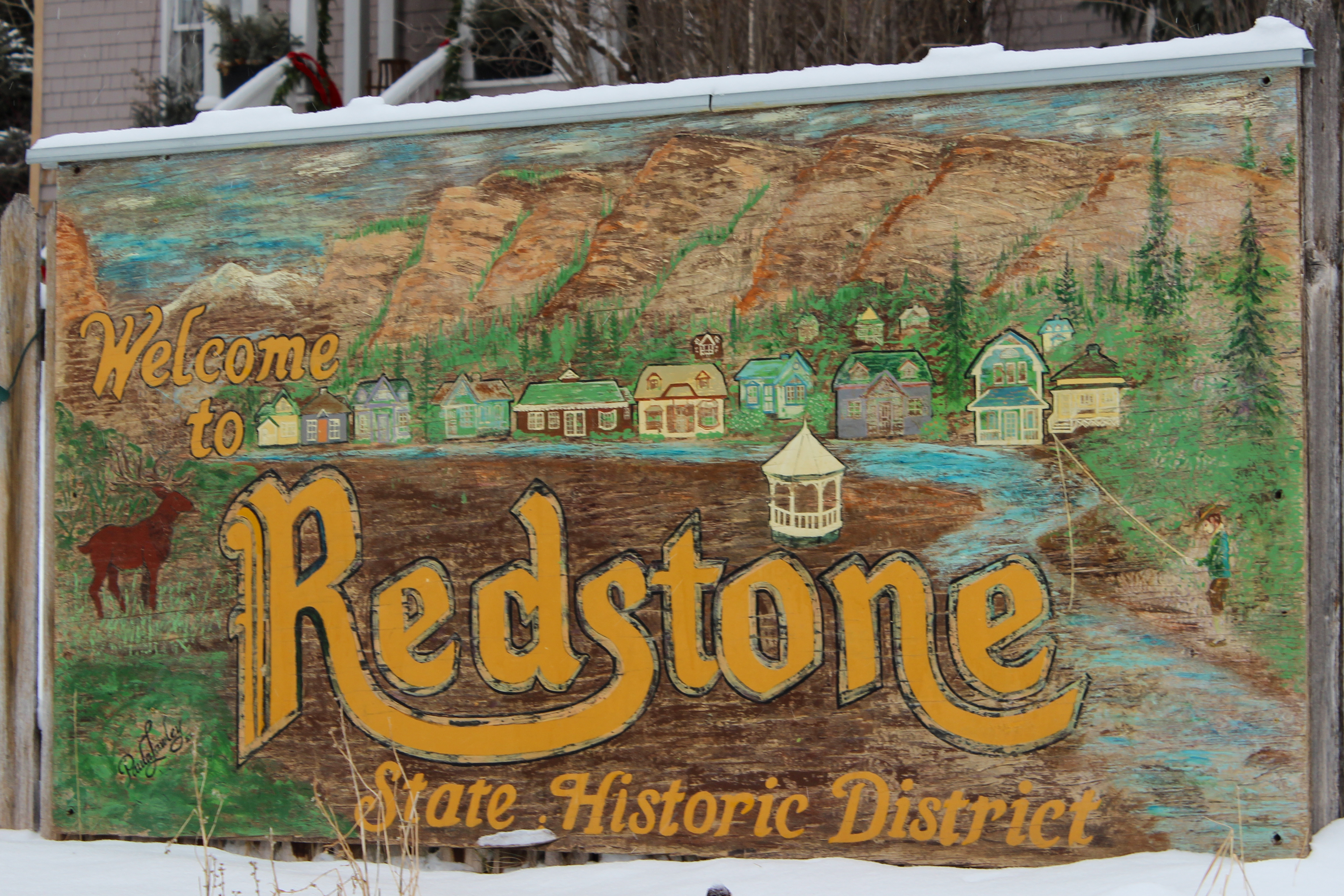 A painted wooden sign, depicting a colorful village, elk, and trees. The signage reads "Welcome to Redstone State Historic District."