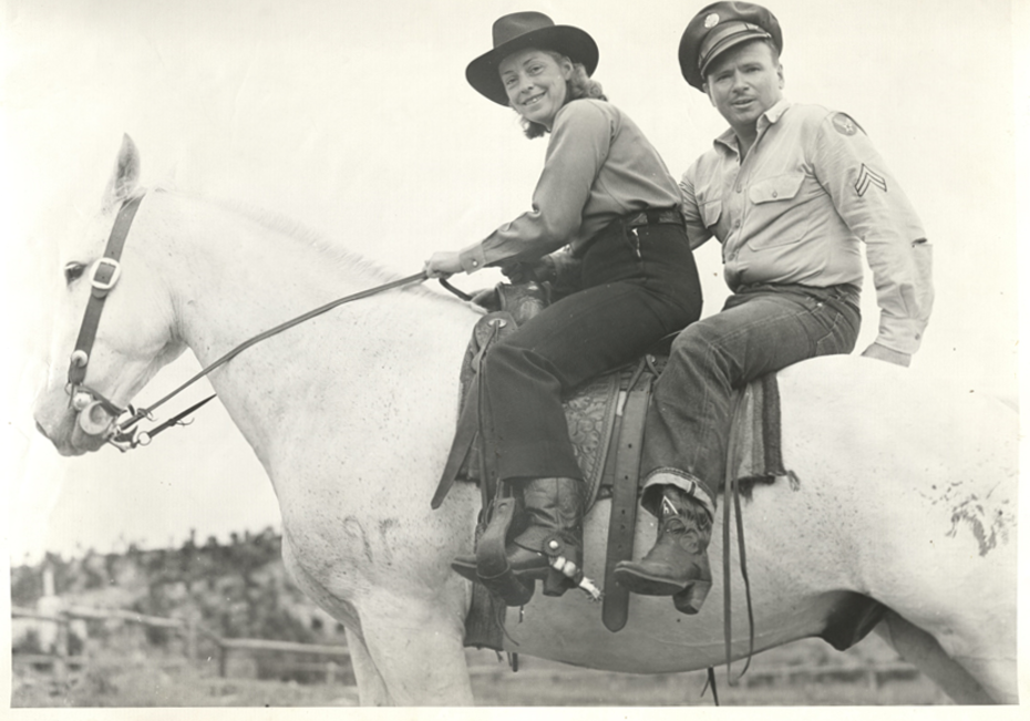 Photo of a woman and a man, riding together on a tall white horse. She wears black pants with black cowboy boots and spurs, and a long-sleeved blouse and a black cowboy hat atop her head of shoulder-length hair. He wear a military uniform shirt and hat, along with denim jeans and cowboy boots. She is holding the horse's reins as they both look at the camera and smile. 